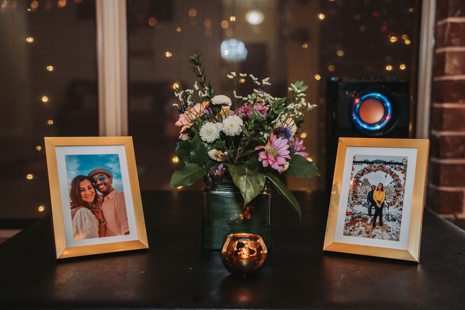 Framed photos and floral centerpiece display on dark table with glowing lights in background.