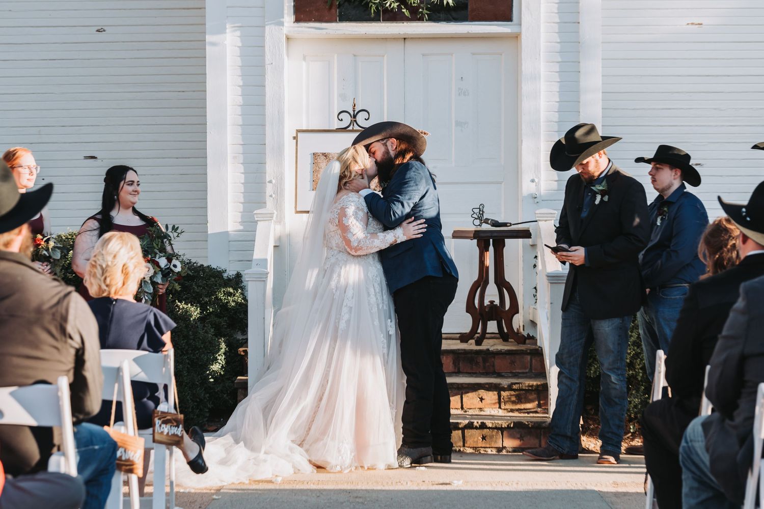 A romantic wedding ceremony kiss under a white gazebo with guests looking on.