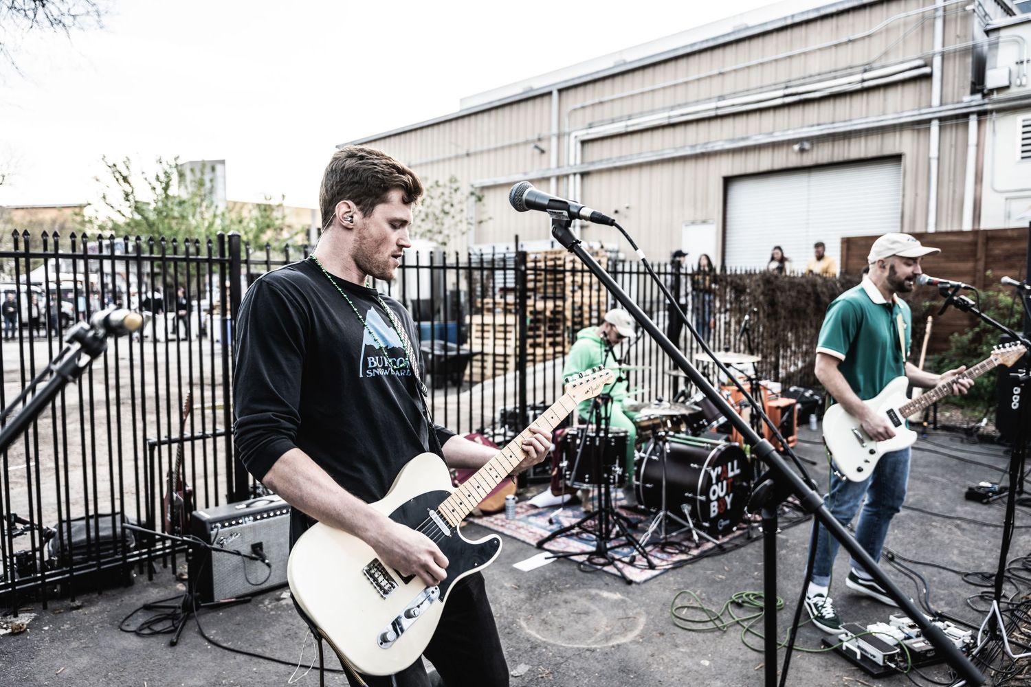 Musicians perform with guitars at outdoor venue behind metal fence.