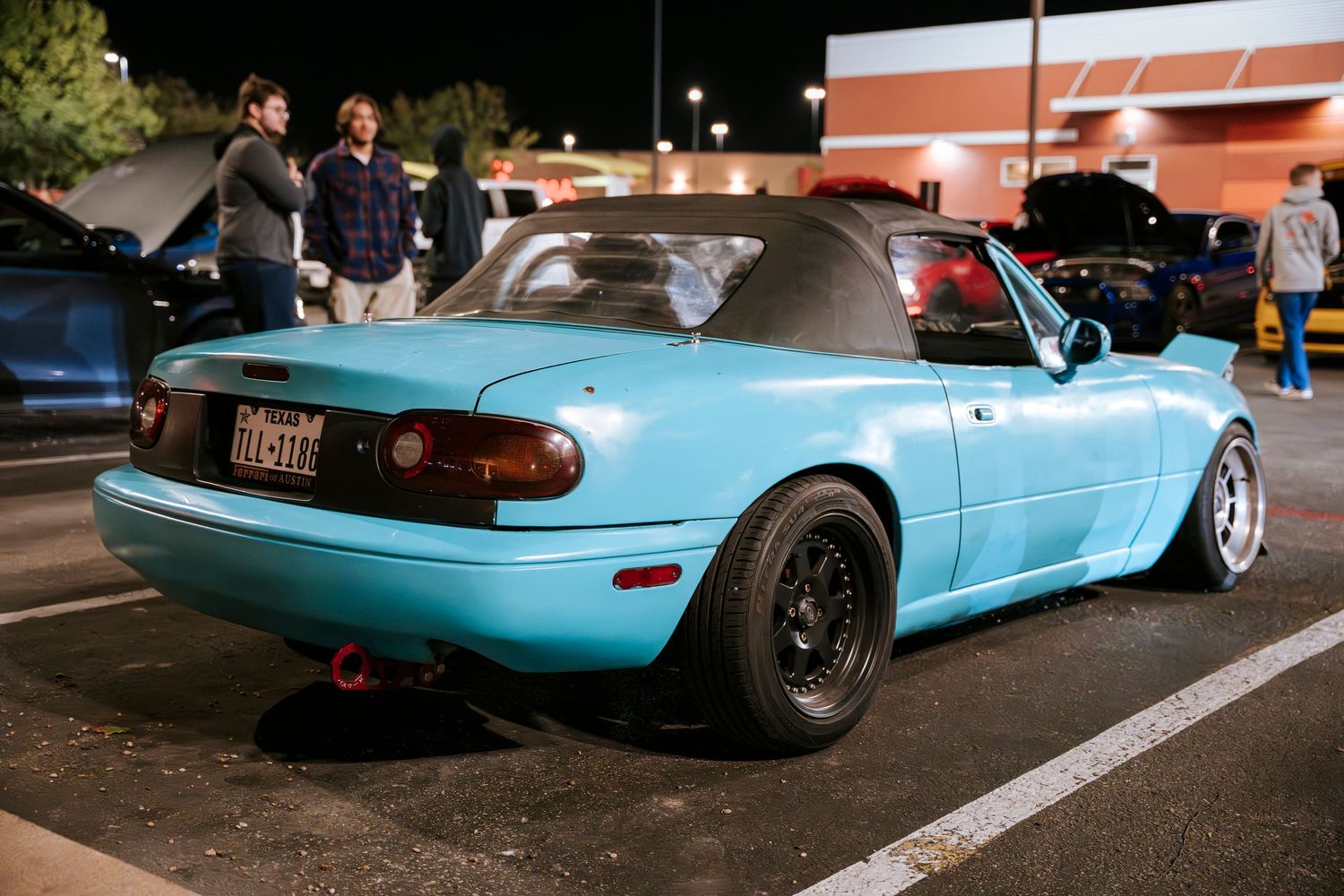 Light blue Mazda Miata convertible with black wheels parked in a nighttime lot.