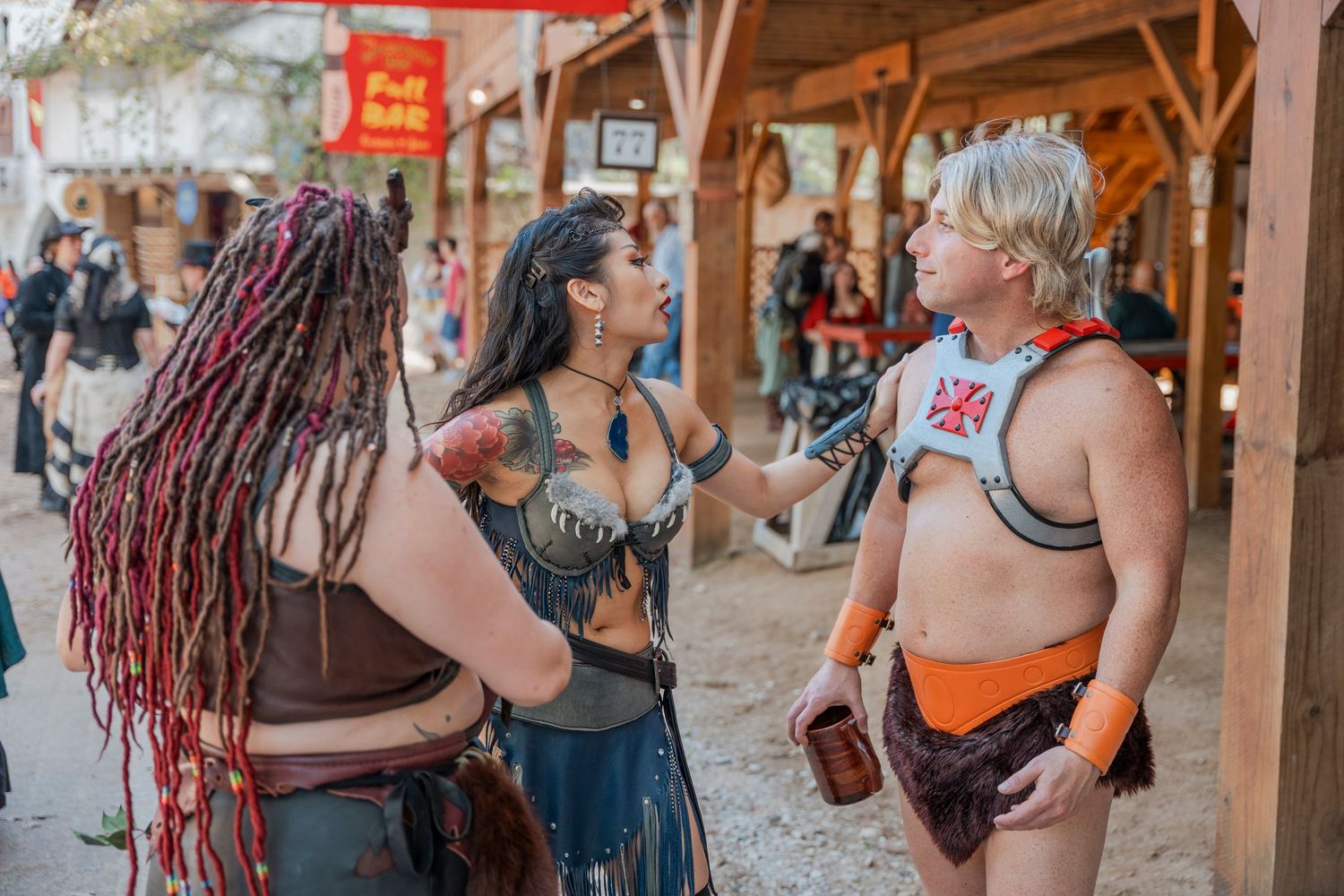 Three people in fantasy costumes converse under wooden pavilions at a renaissance festival.