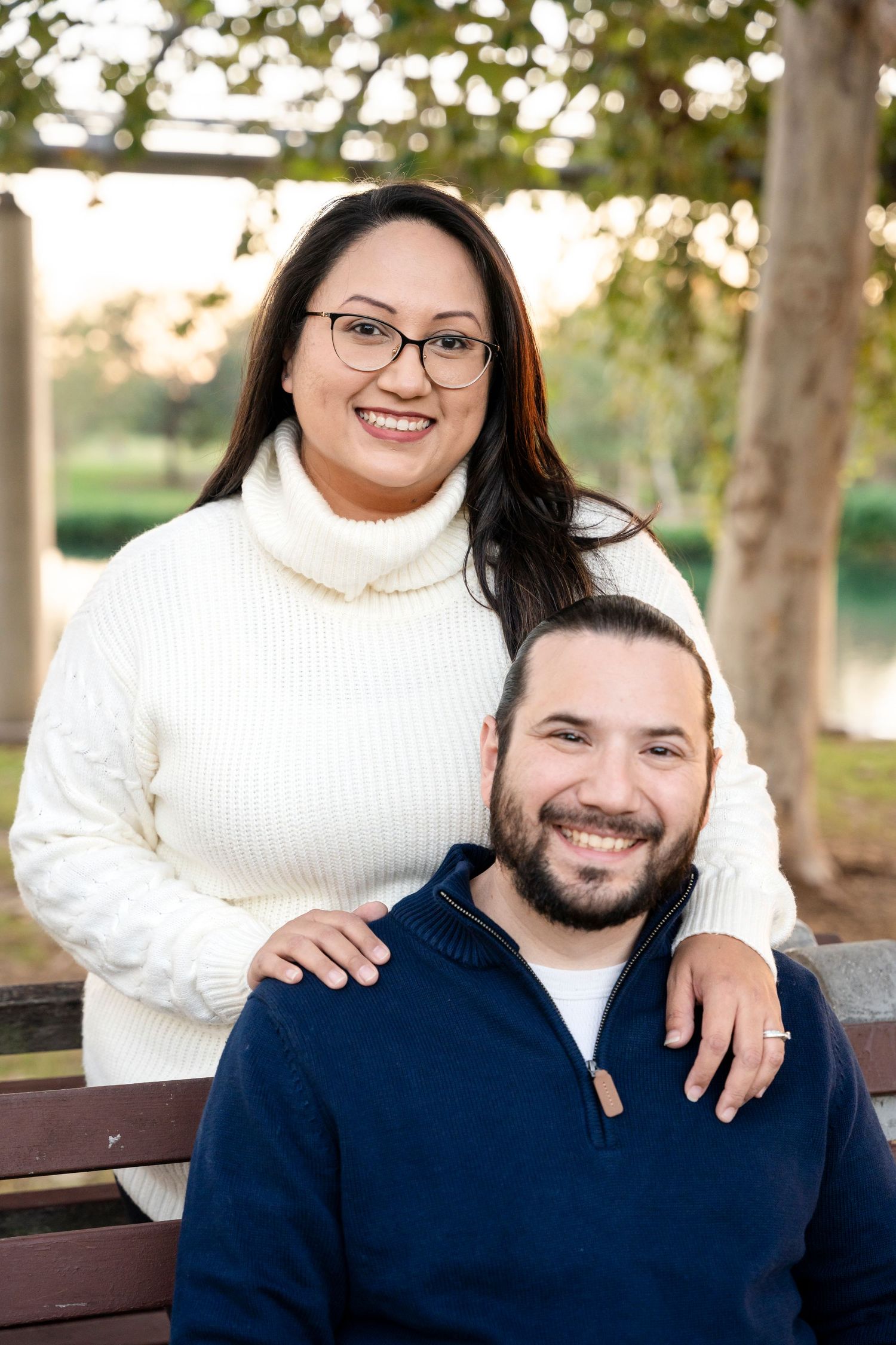 Couple in coordinating sweaters pose on park bench during autumn sunset.