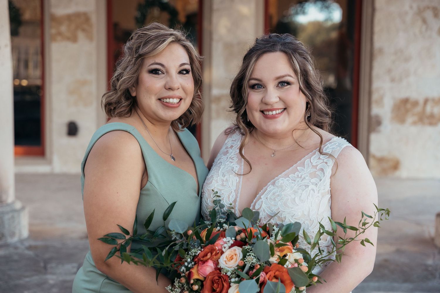 Two attendants pose with beautiful fall-themed bouquets on a stone patio.