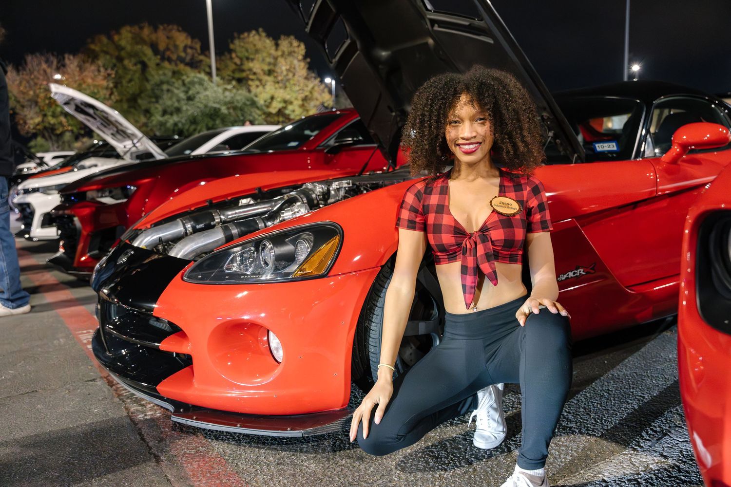 Person in red tied top and jeans sits casually on red car in outdoor lot.