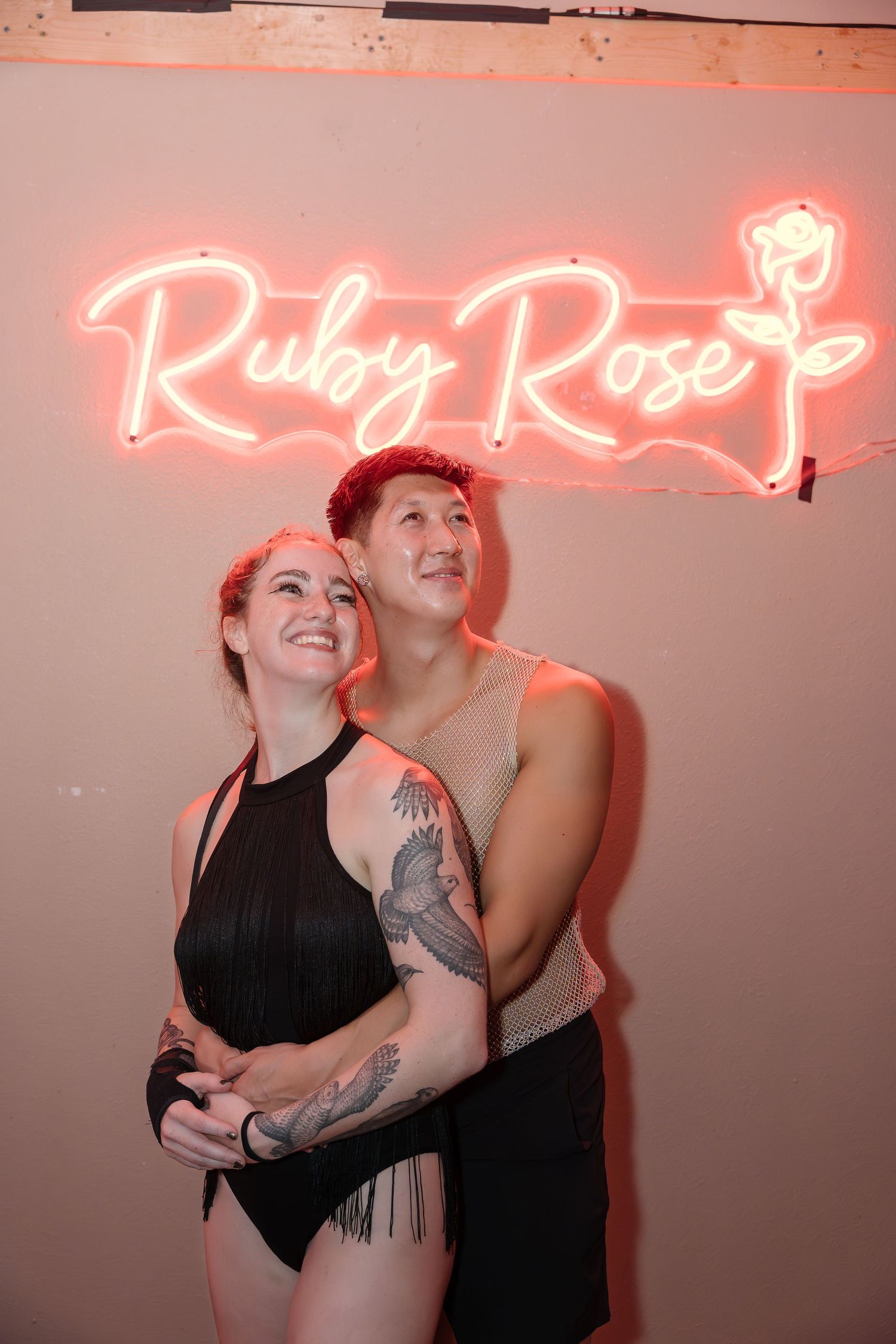 Two performers pose playfully beneath a pink neon 'Ruby Rose' sign in a dimly lit venue.