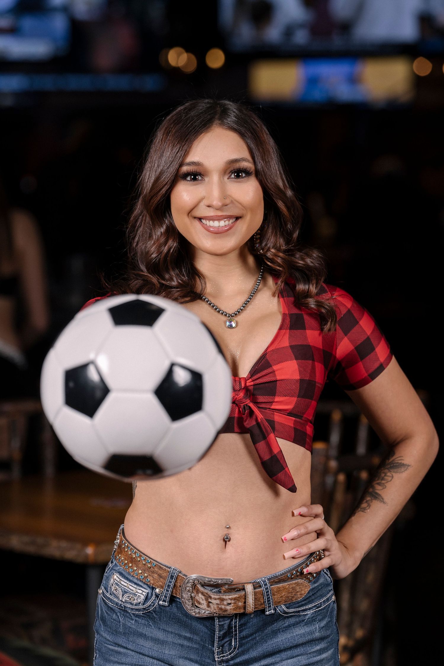 Woman in buffalo plaid crop top poses with soccer ball at a sports bar.