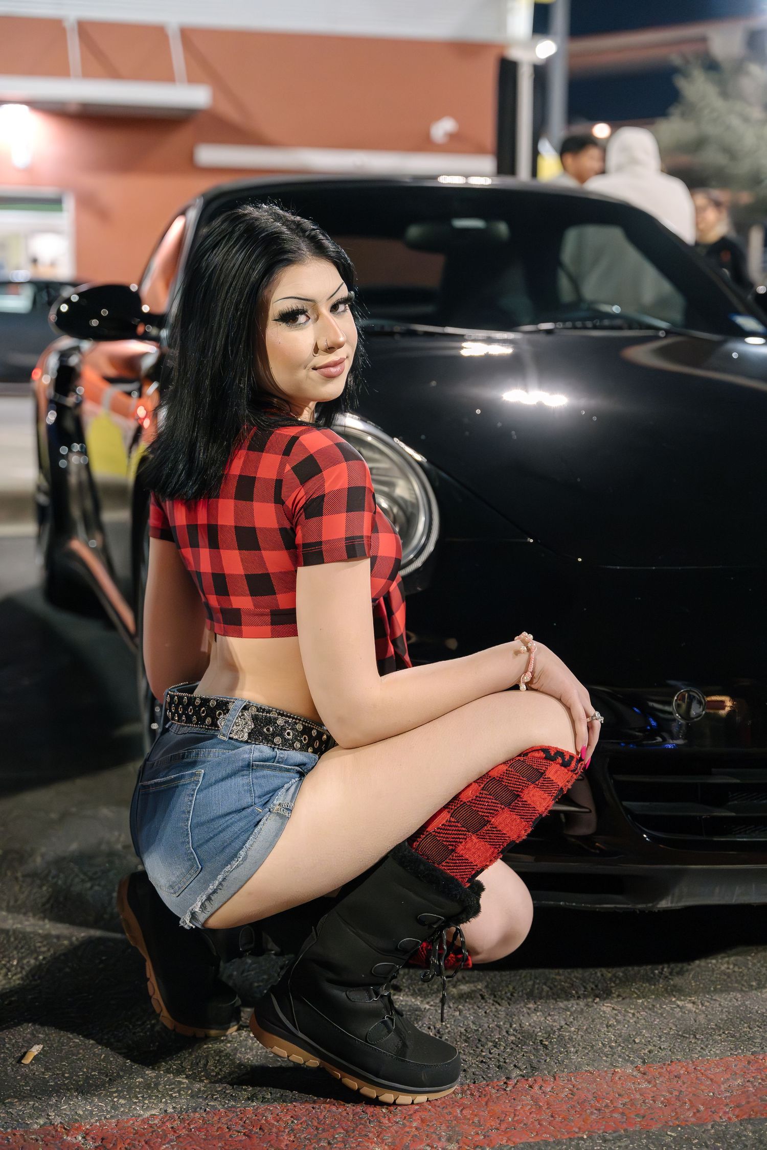 Woman in plaid crop top crouching next to black sports car at night.