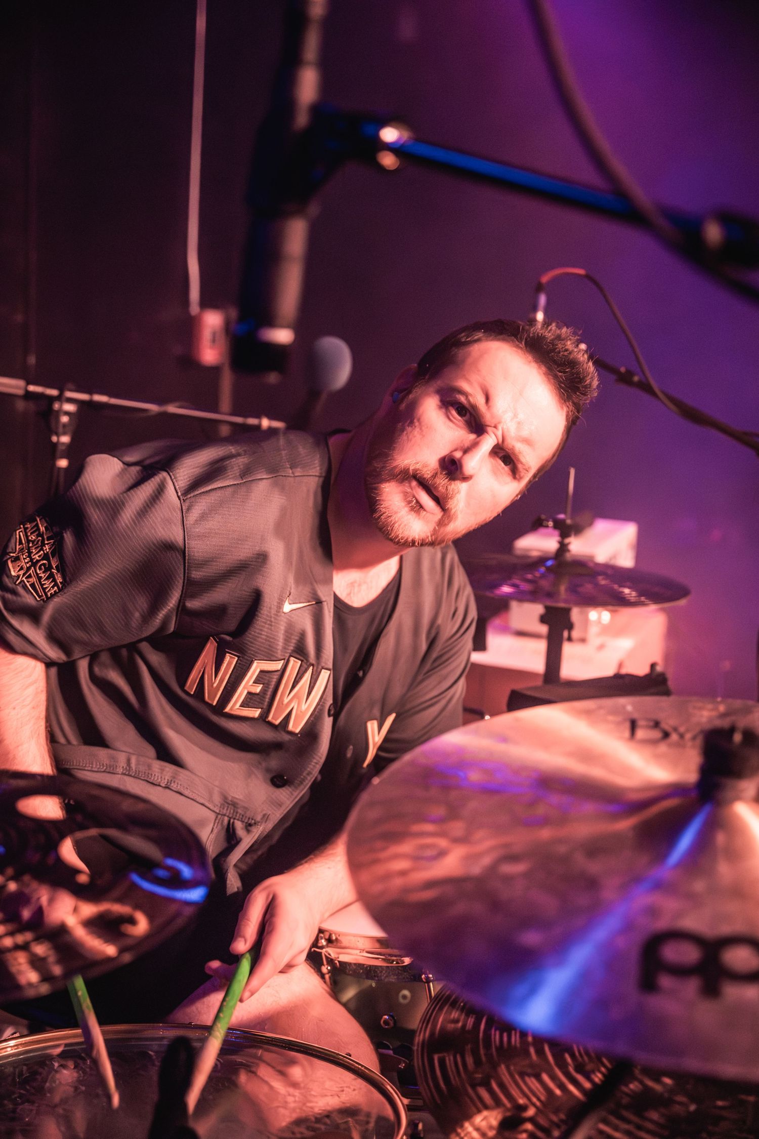 A drummer in a grey sweatshirt performs passionately under purple stage lighting.