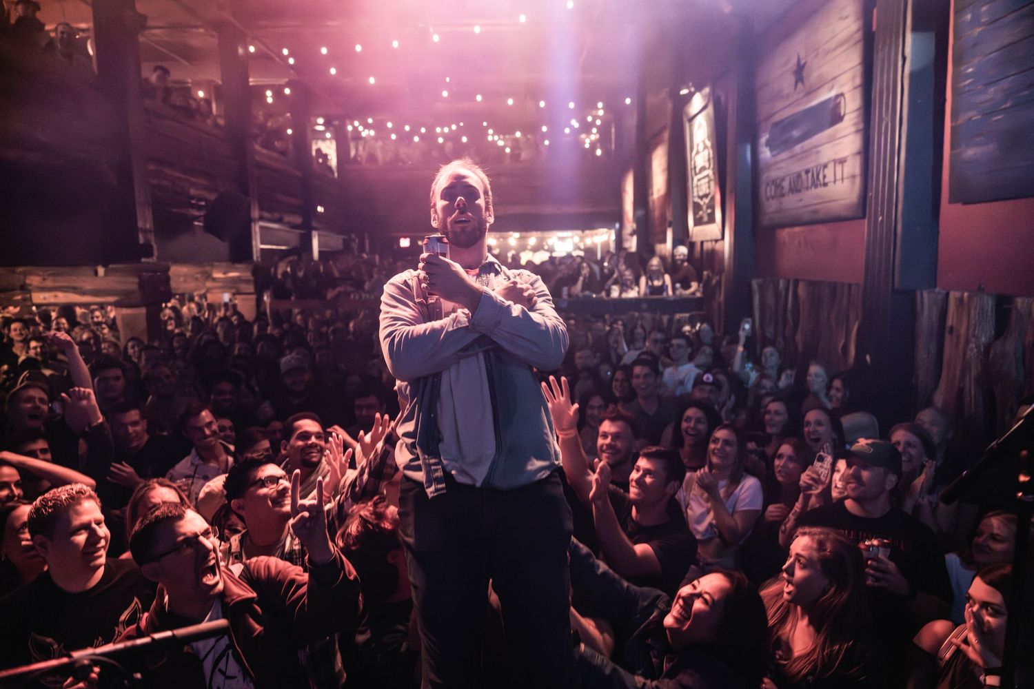 Performer on stage surrounded by enthusiastic crowd in dark venue with purple lighting.
