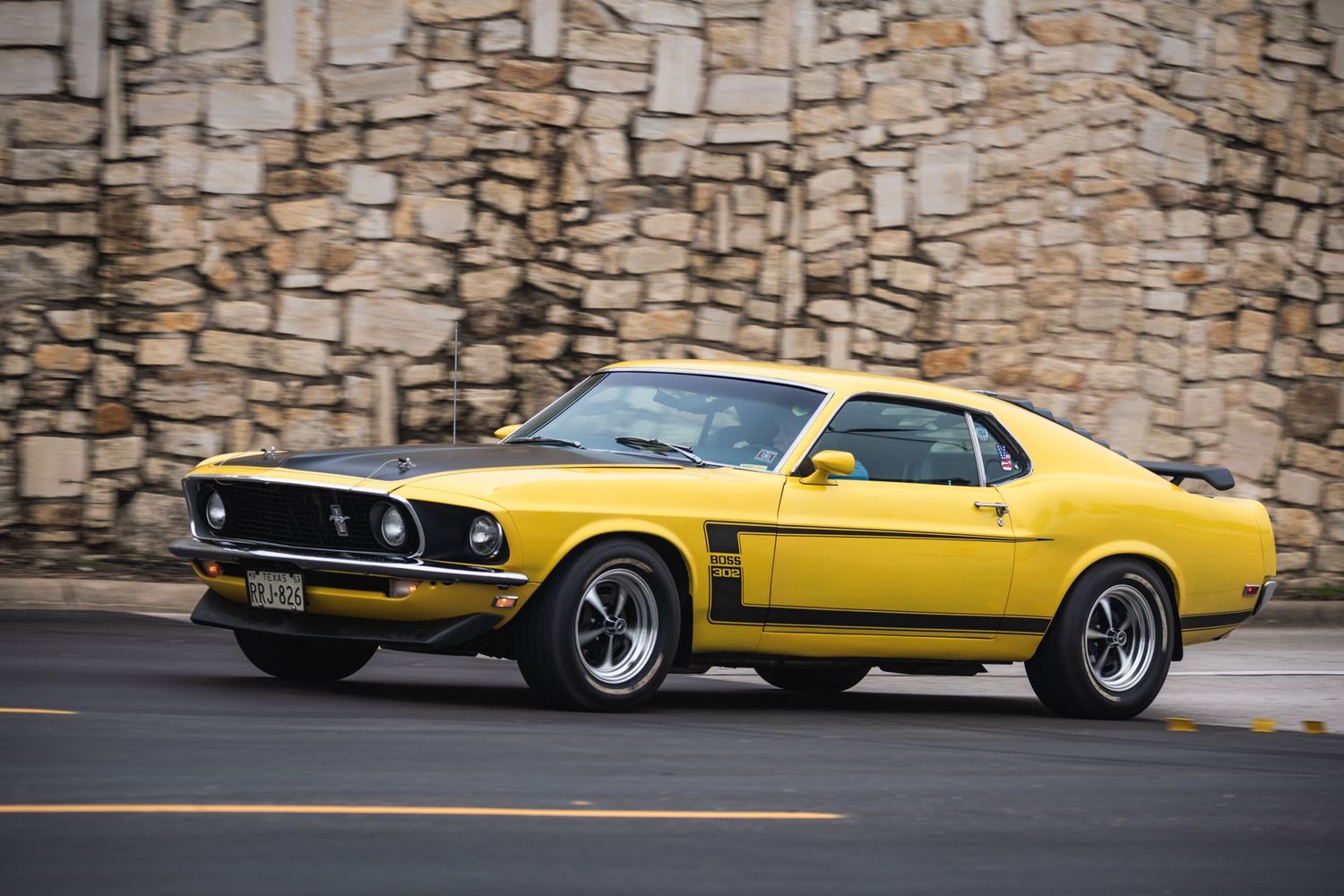 Yellow 1969 Ford Mustang Boss parked on street against stone wall backdrop.