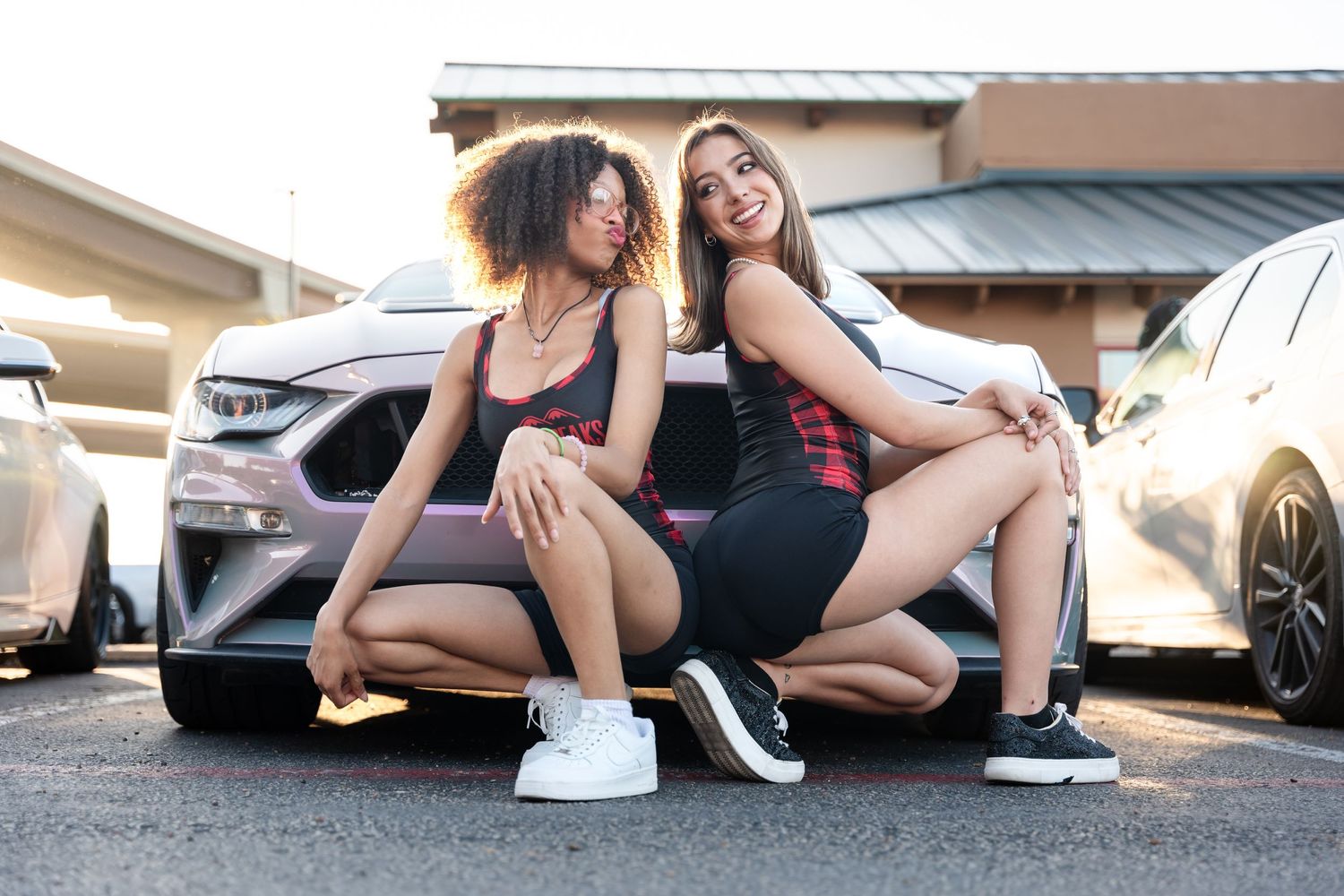 Two friends posing playfully next to a parked car in a parking lot during sunset.