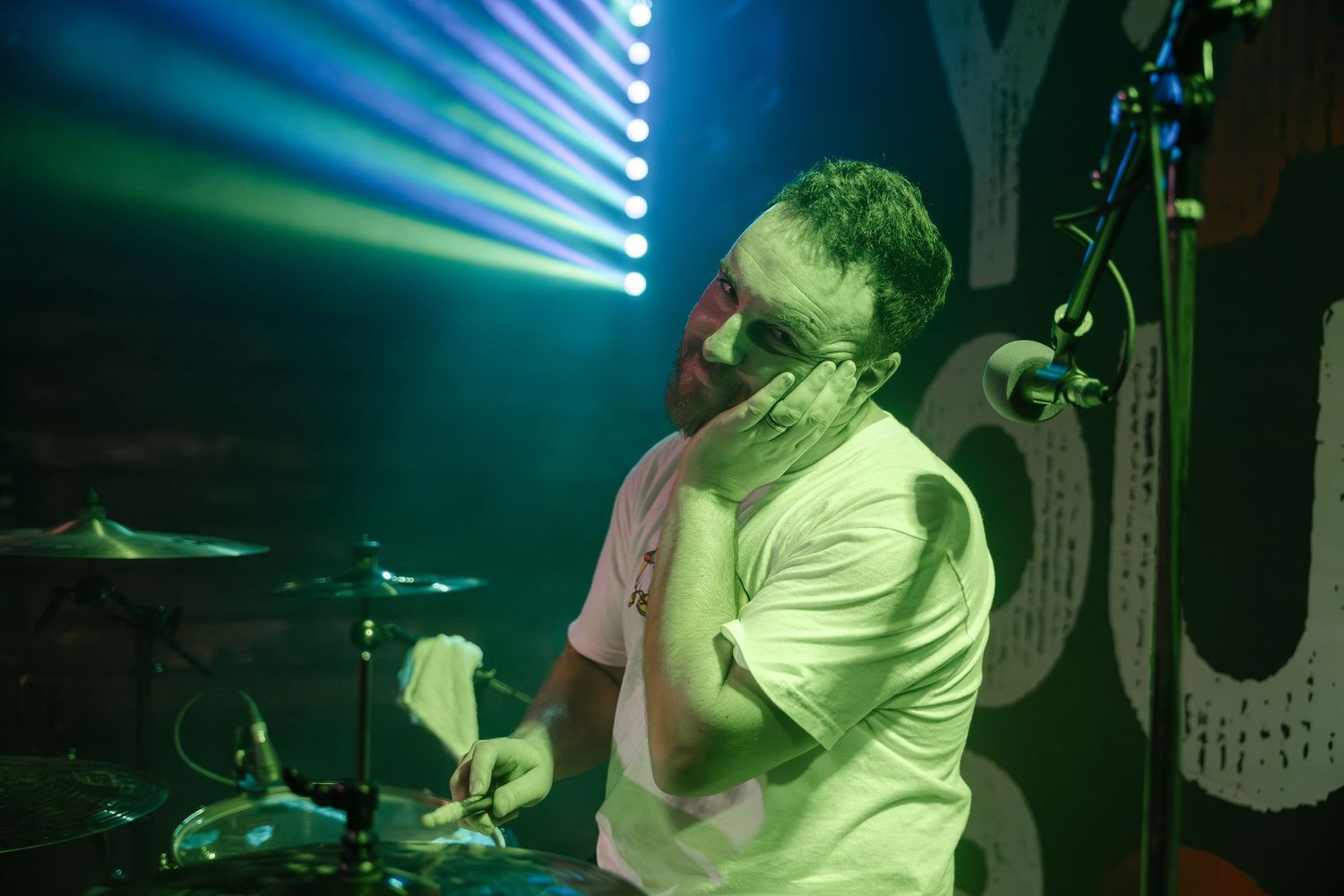 Performer singing into microphone under green stage lights with drum kit visible.