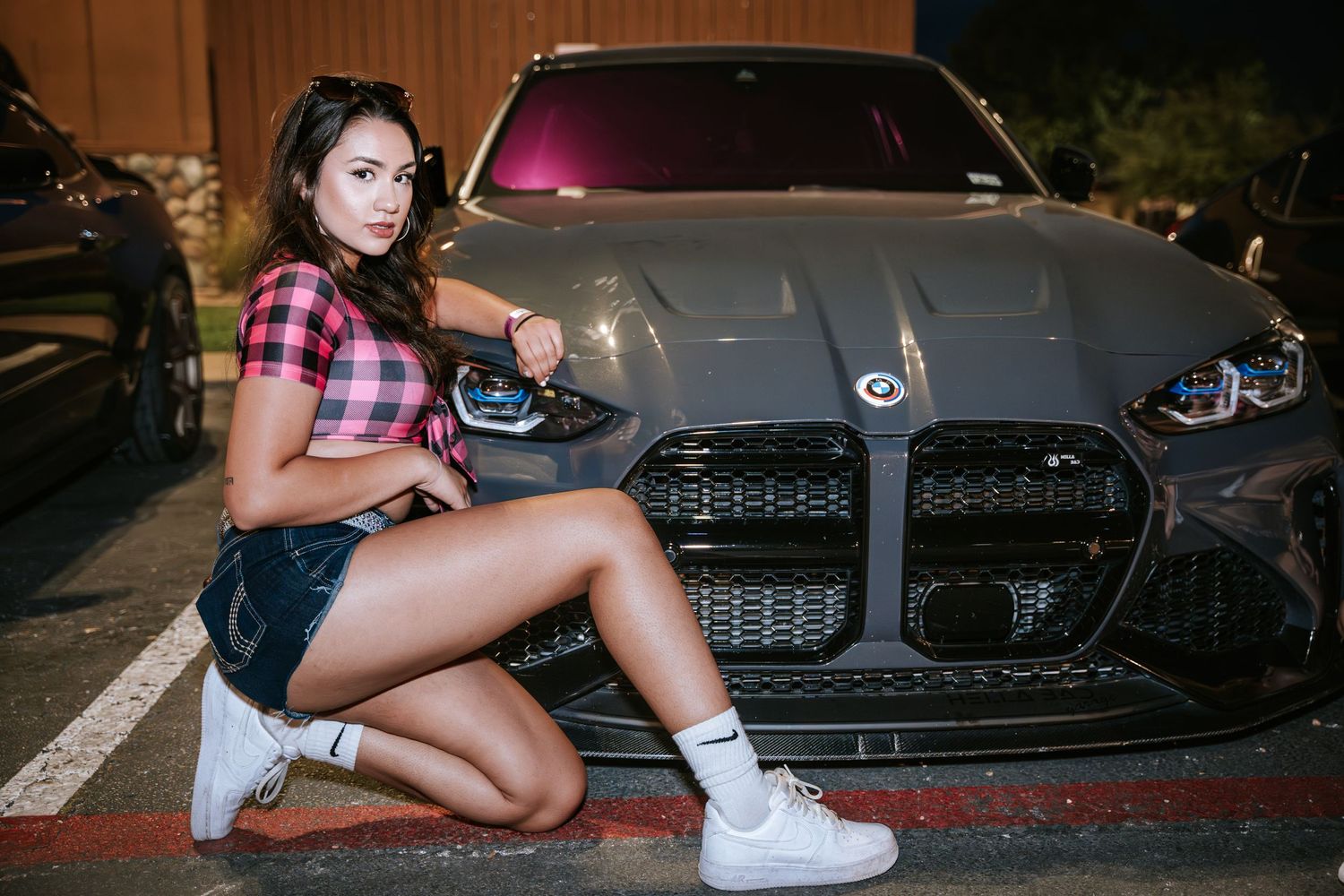 Person in plaid crop top posing with gray BMW M4 in parking lot at night.