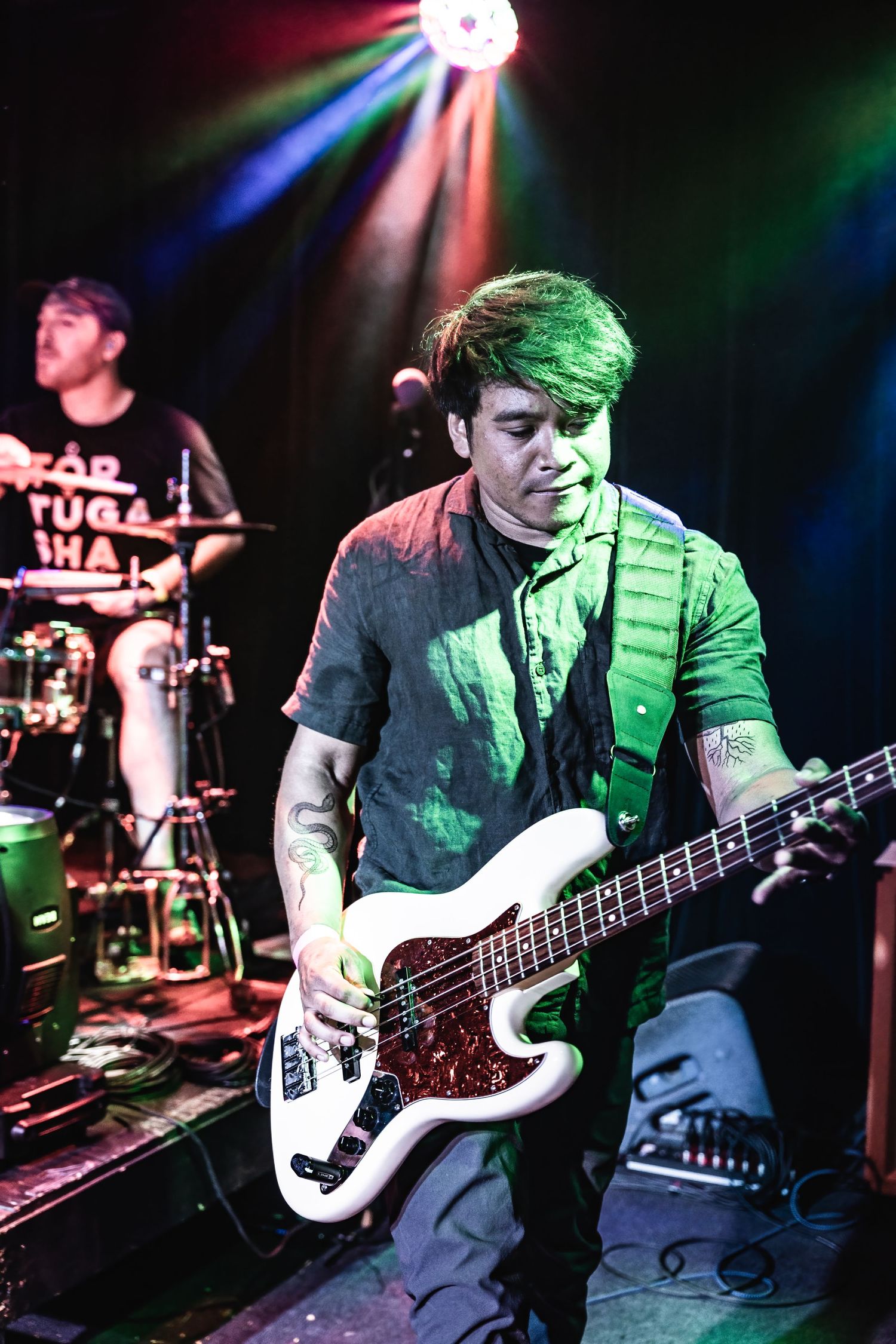 Bass player performs under colorful stage lighting while drummer plays in background.