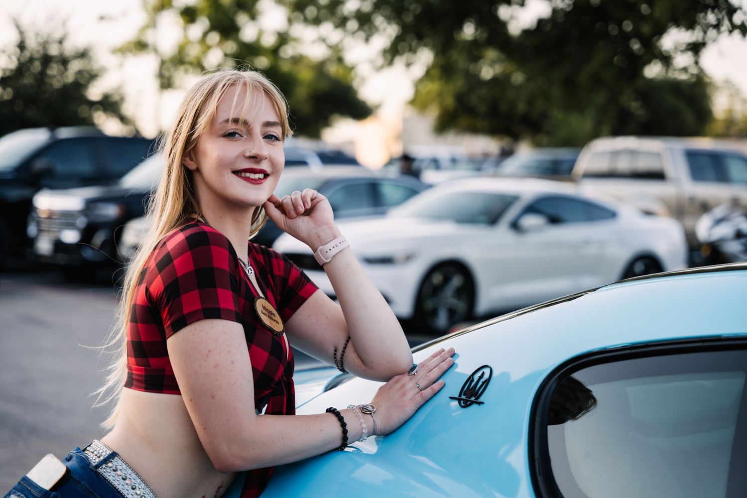 Person in casual summer outfit smiles while leaning on a blue car at sunset.