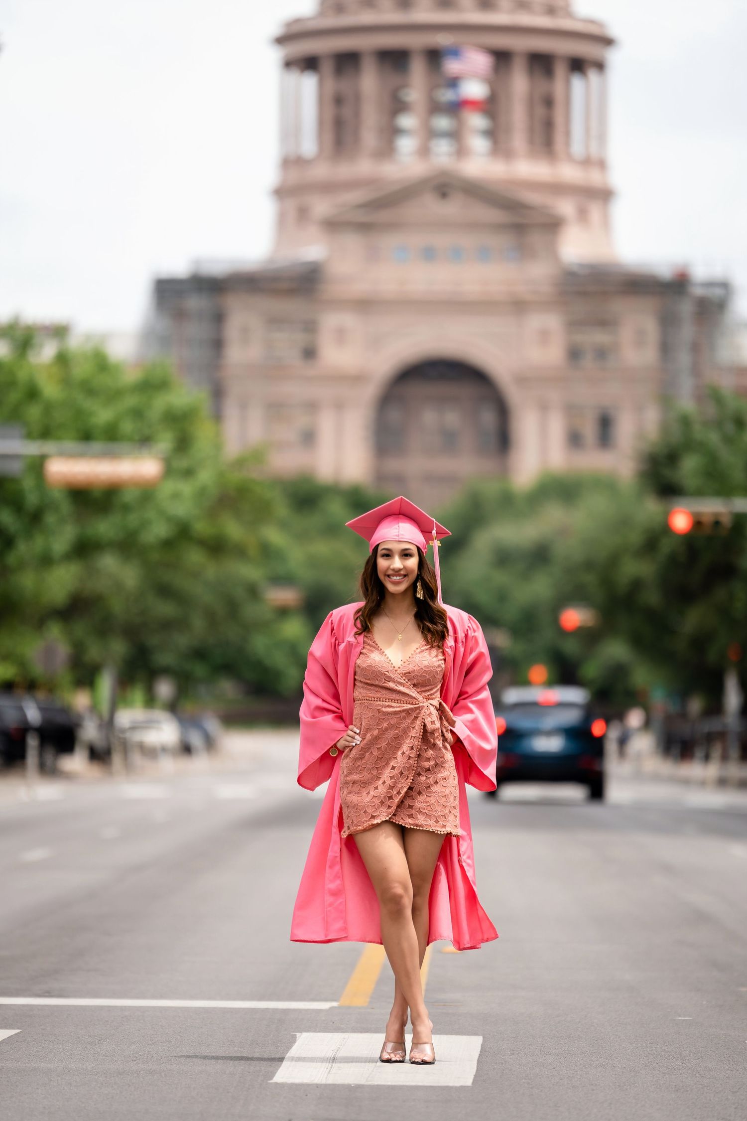 Graduate in pink attire strikes elegant pose with Texas State Capitol in background.