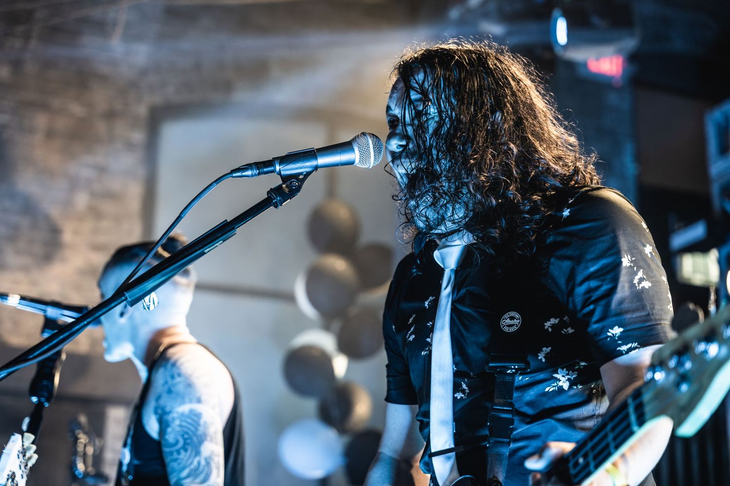 A performer with long hair plays guitar and sings into a microphone on a dimly lit stage.