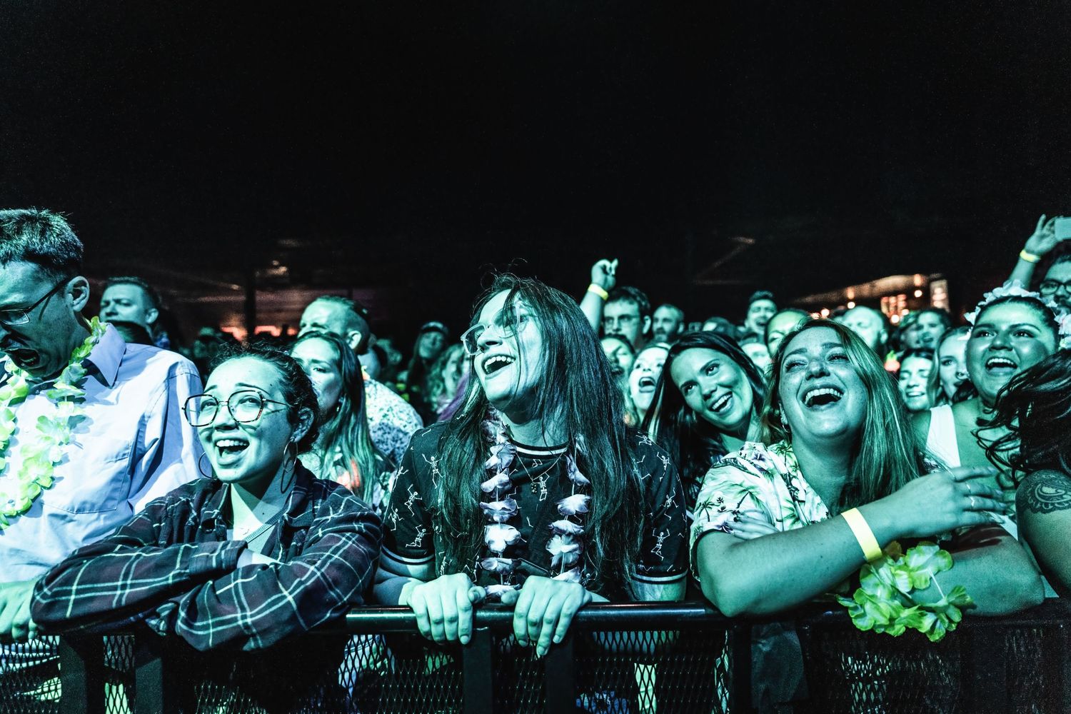 Excited concert crowd bathed in blue lighting lean against security barrier while cheering.