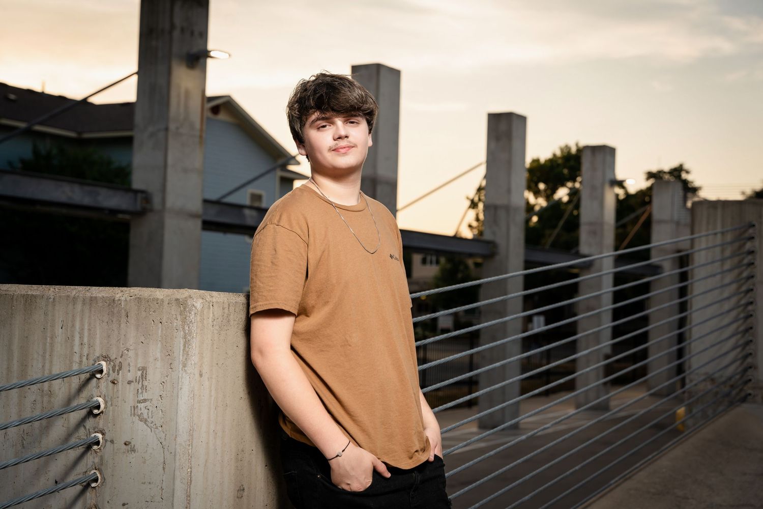 Someone in a brown t-shirt poses against an urban backdrop at sunset.