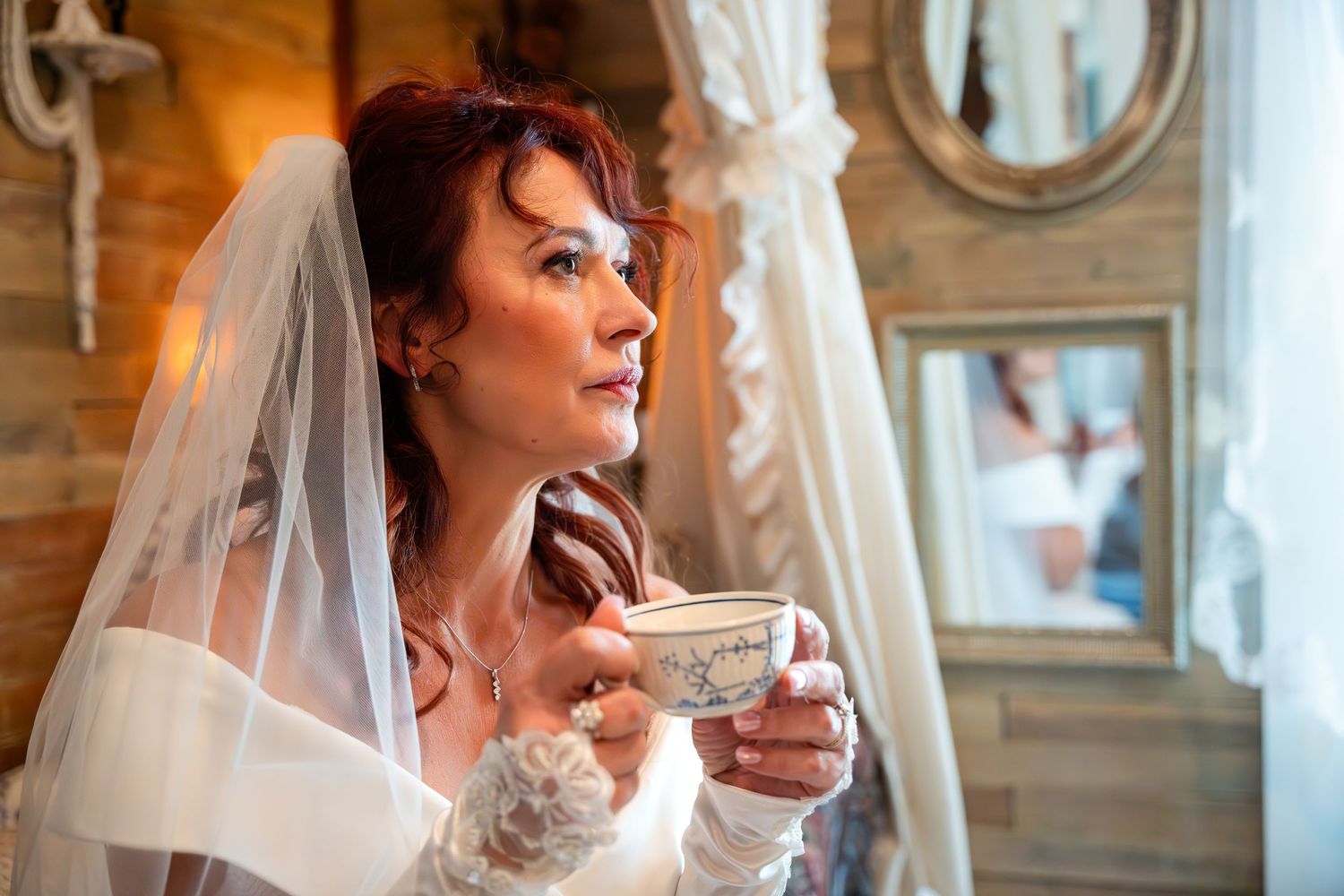Bride in white lace gown and veil takes a quiet moment with teacup during wedding preparations.