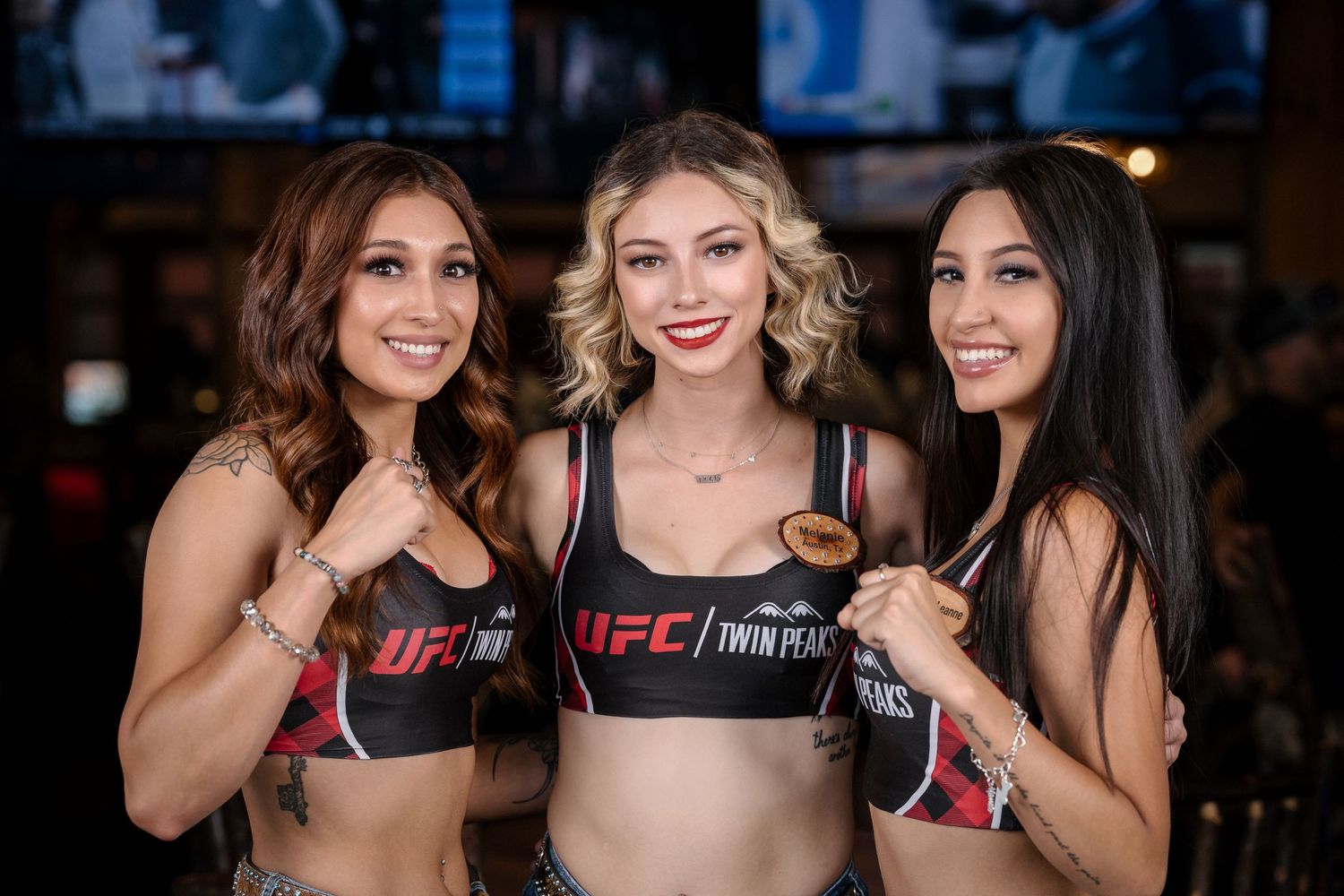Three models wearing UFC promotional gear pose with boxing gloves at sports venue.