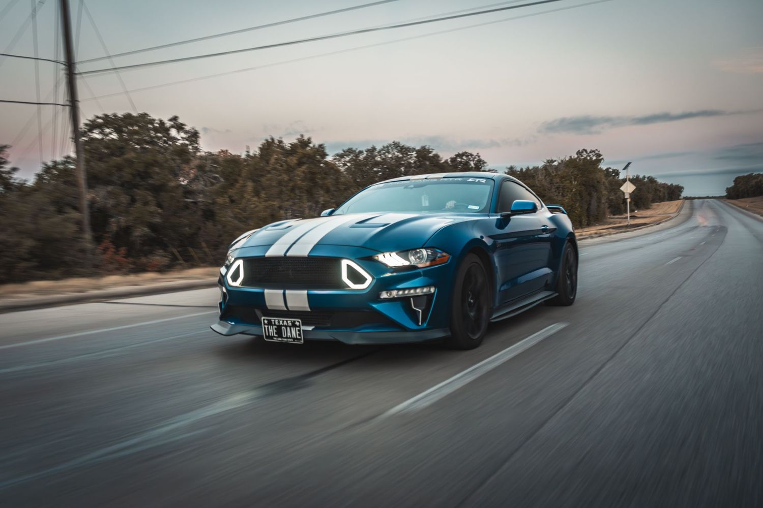 Blue Ford Mustang with dual white racing stripes cruising on a curved road at dusk.