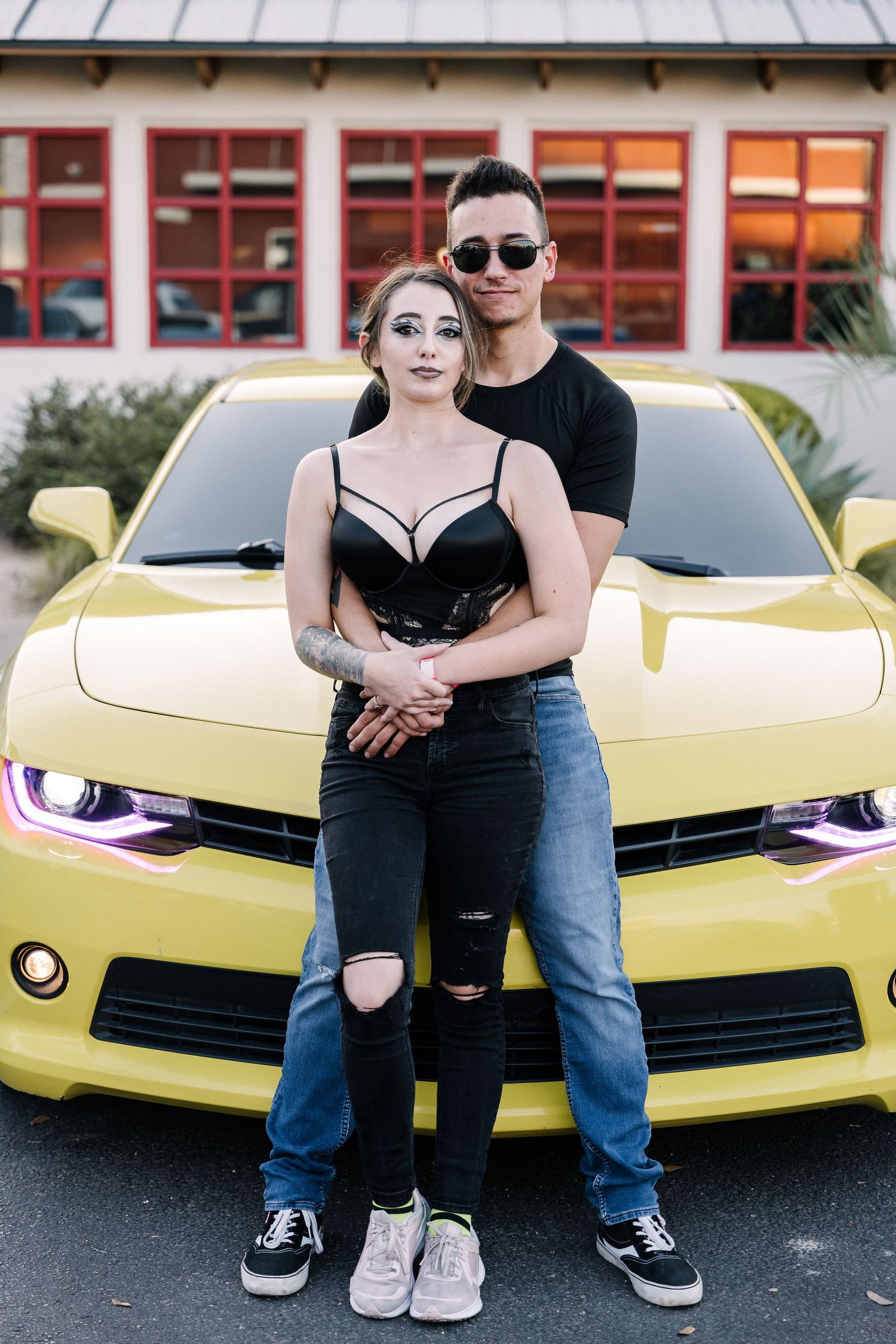 Couple poses together against yellow Chevrolet Camaro at sunset car meet.