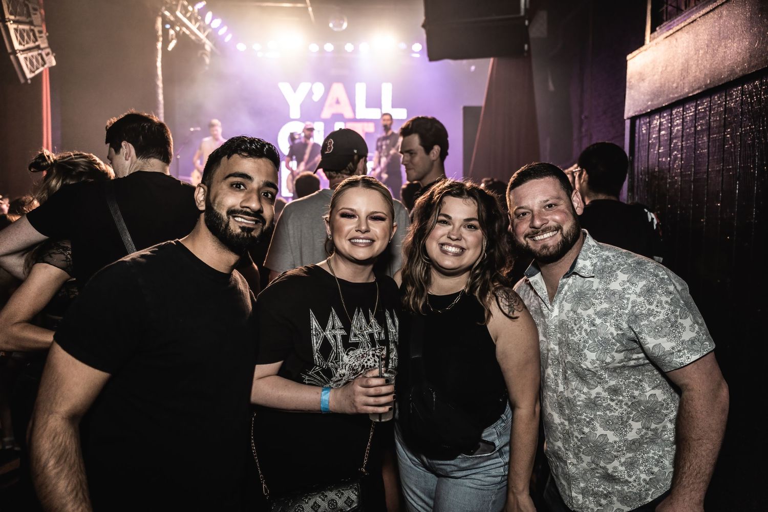 Concert attendees enjoying a night out at a live music event with Y'all Out Boy signage in the background.