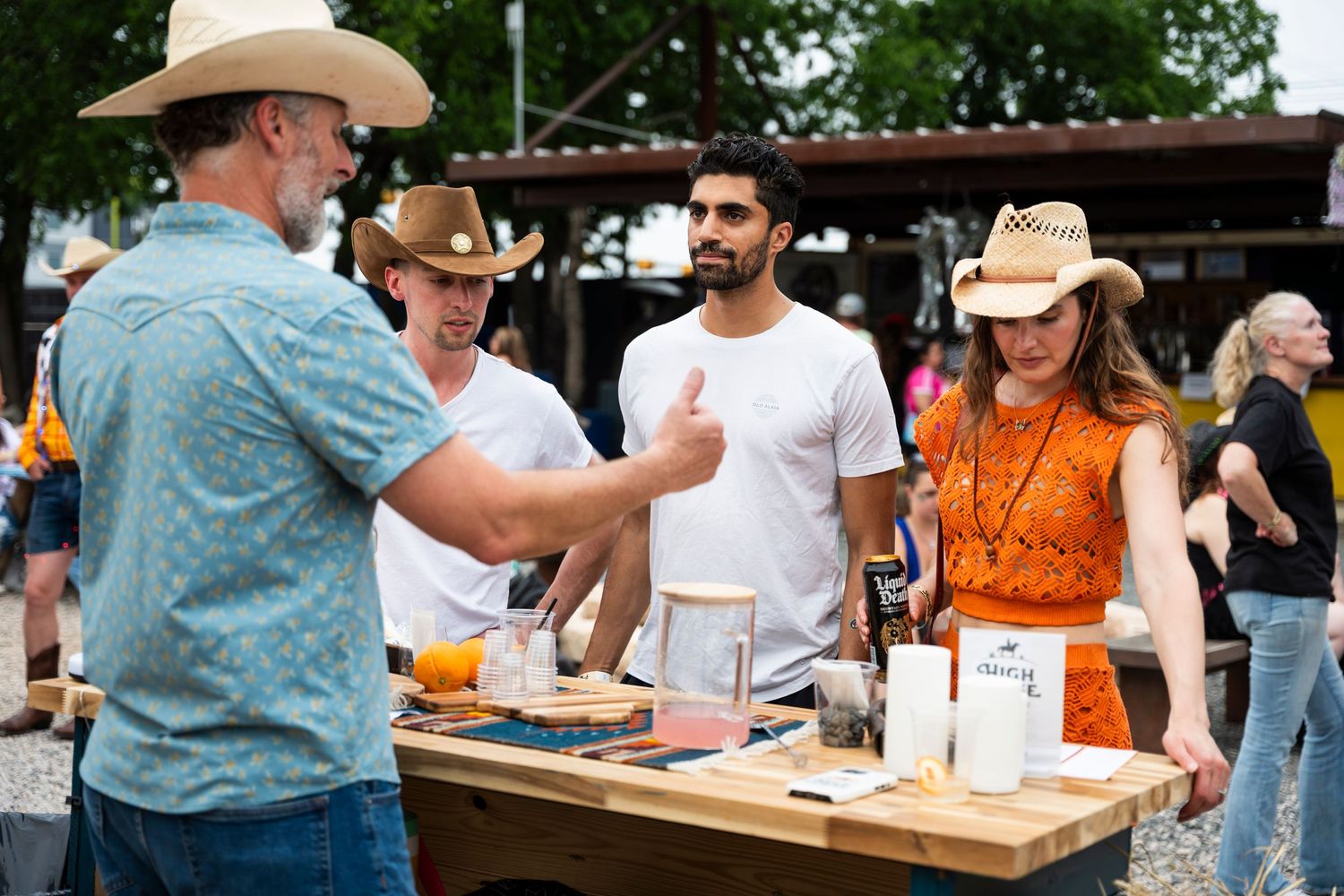 People enjoying a sunny outdoor event with vendors selling goods at wooden tables while wearing cowboy hats.