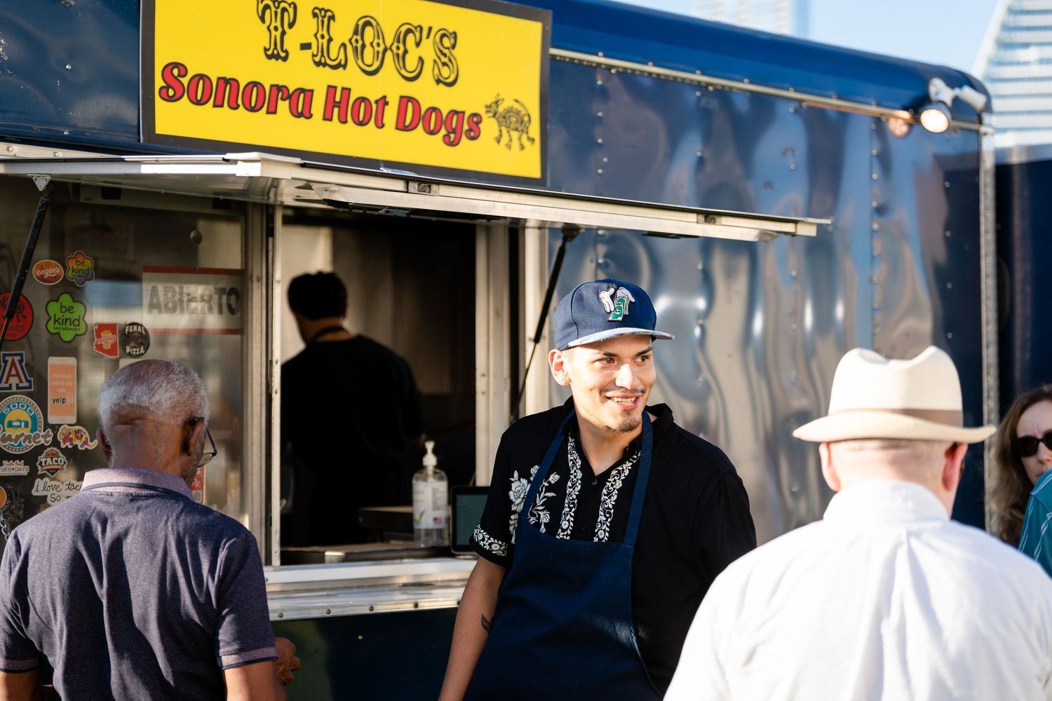 Food truck customers line up at Sonora Hot Dogs vendor cart with city buildings in background.