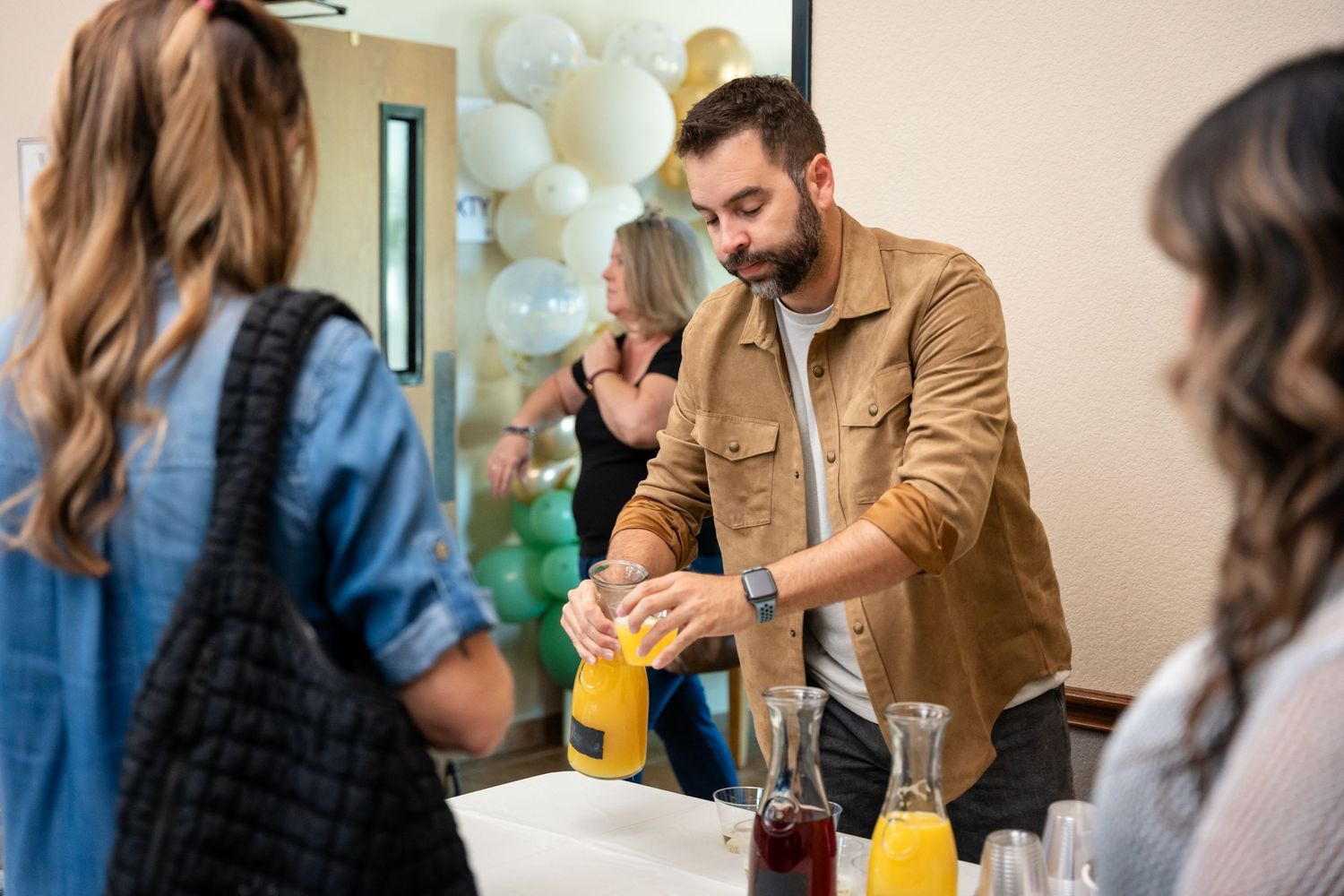 A person in a brown jacket pouring drinks at a party with decorative balloons.