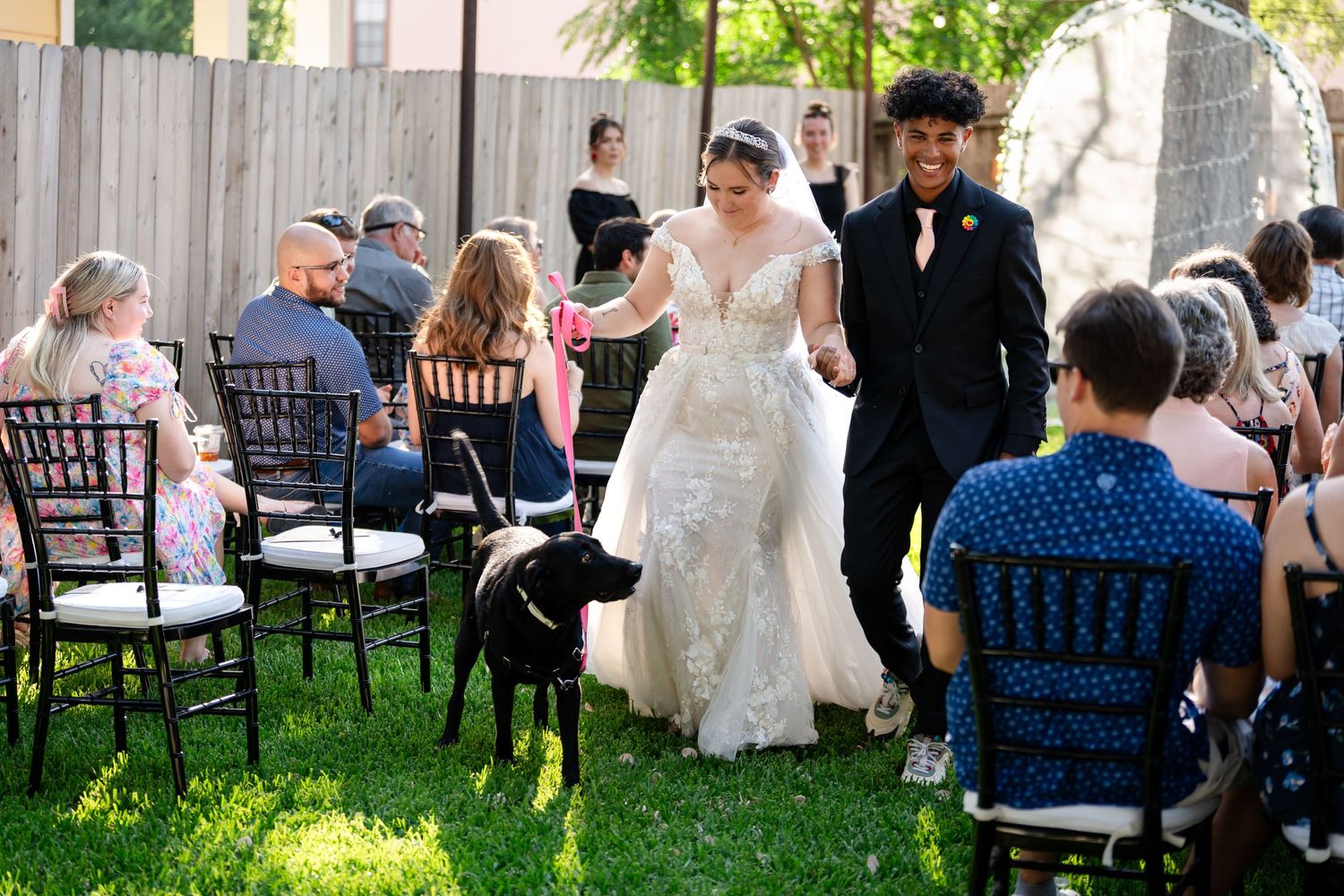 A couple walks down the aisle with their dog during an outdoor garden ceremony.