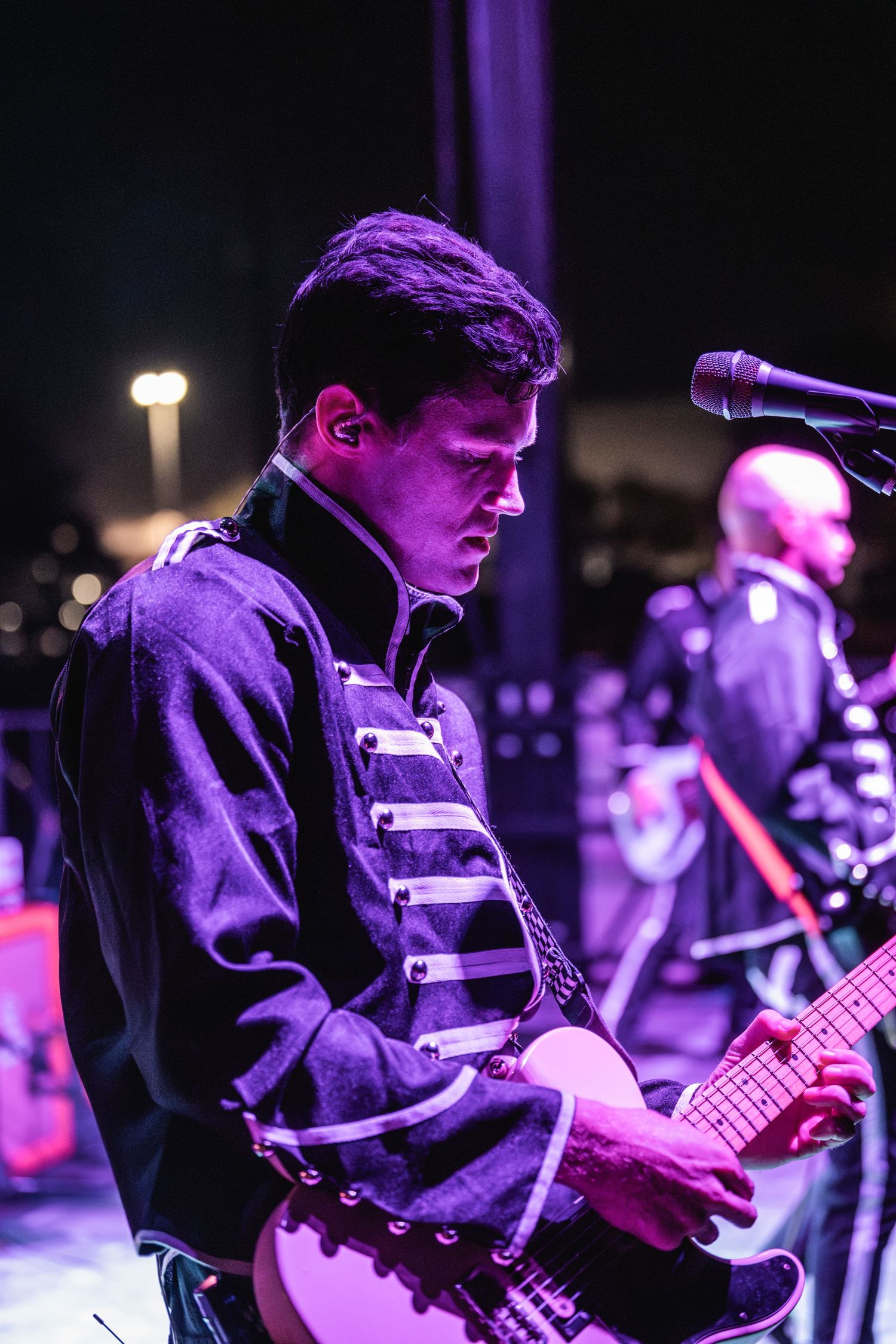 Guitarist performing on stage wearing a military-style jacket under dramatic purple lighting.