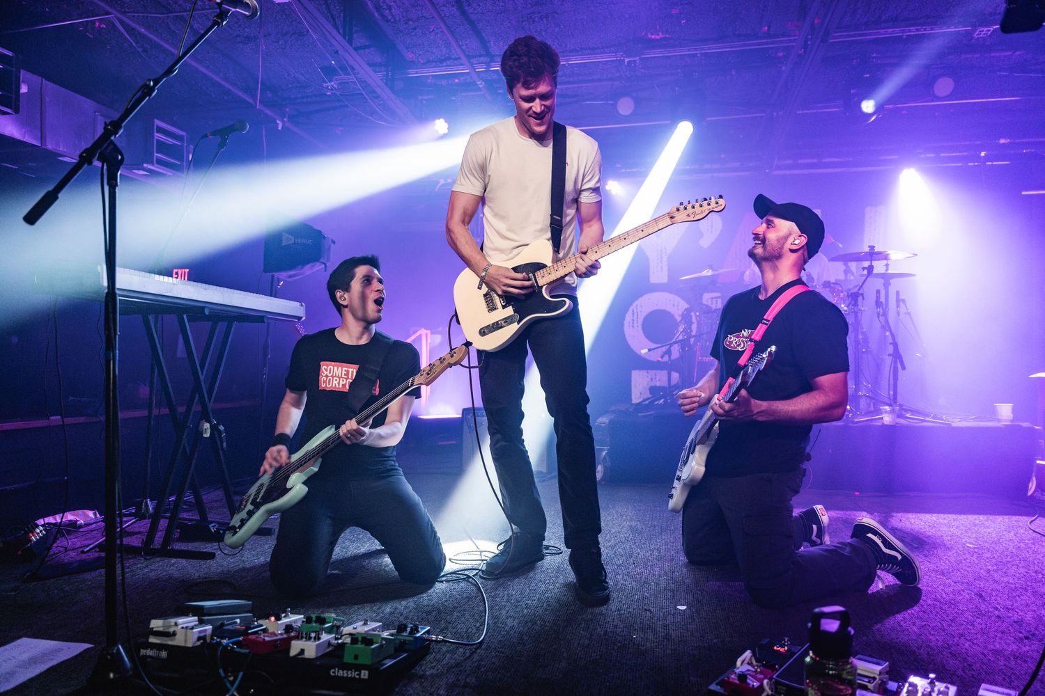 Three musicians performing energetically on stage with electric guitars under dramatic blue lighting and fog effects.