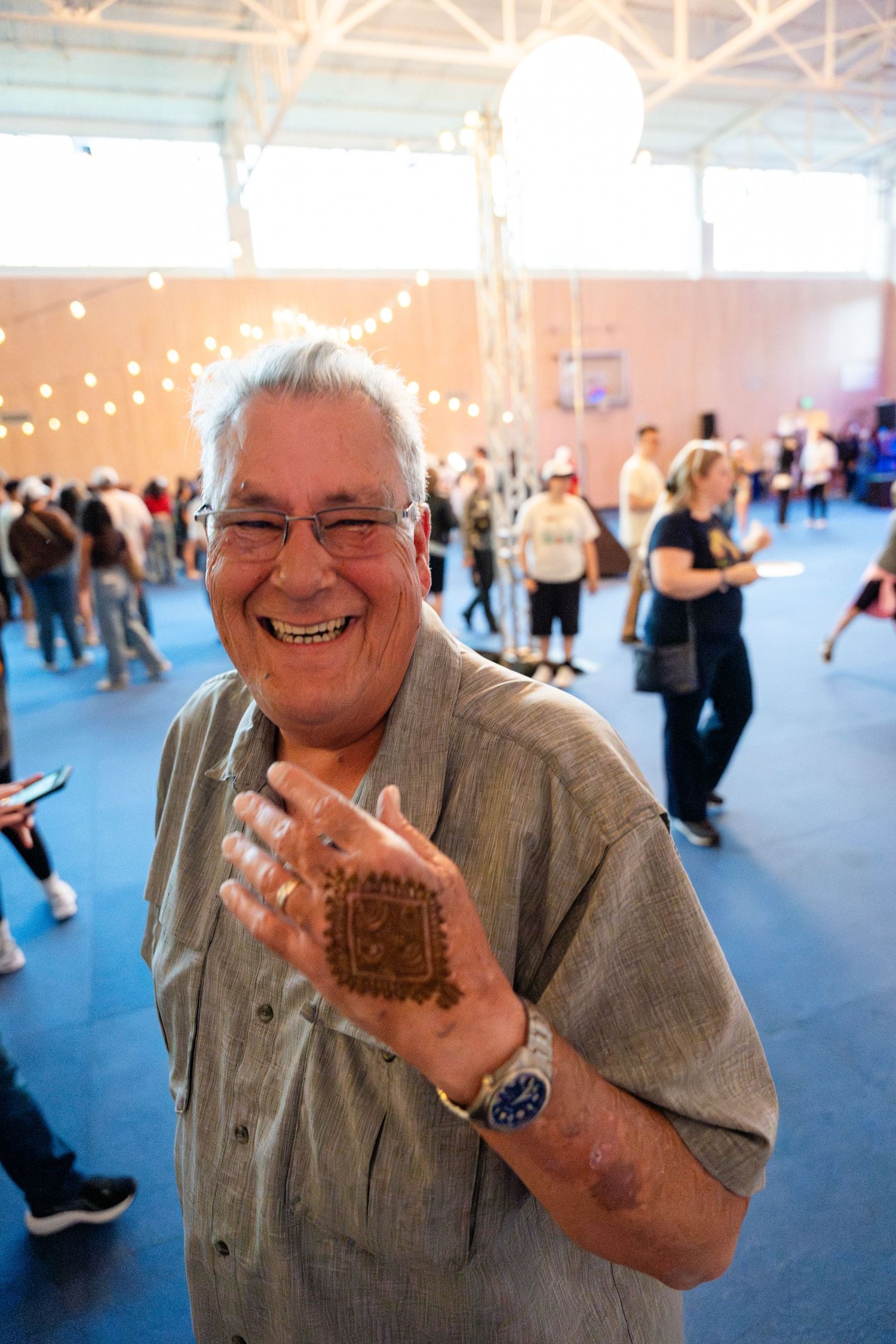 A person wearing glasses shows off decorative henna art on their hand at an indoor event with string lights and people dancing.