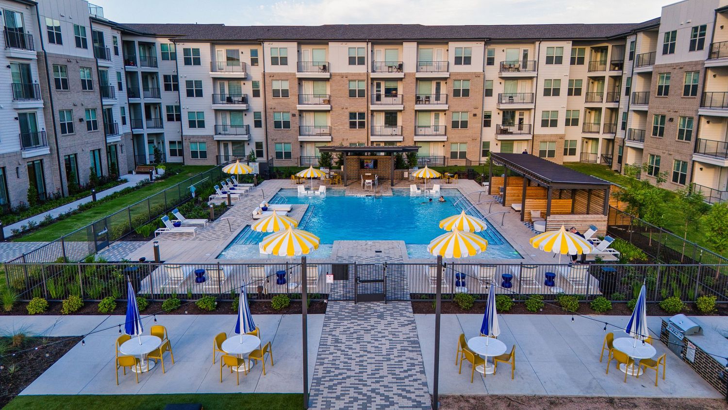 Aerial view of apartment complex pool area with yellow umbrellas and wooden pergolas at dusk.