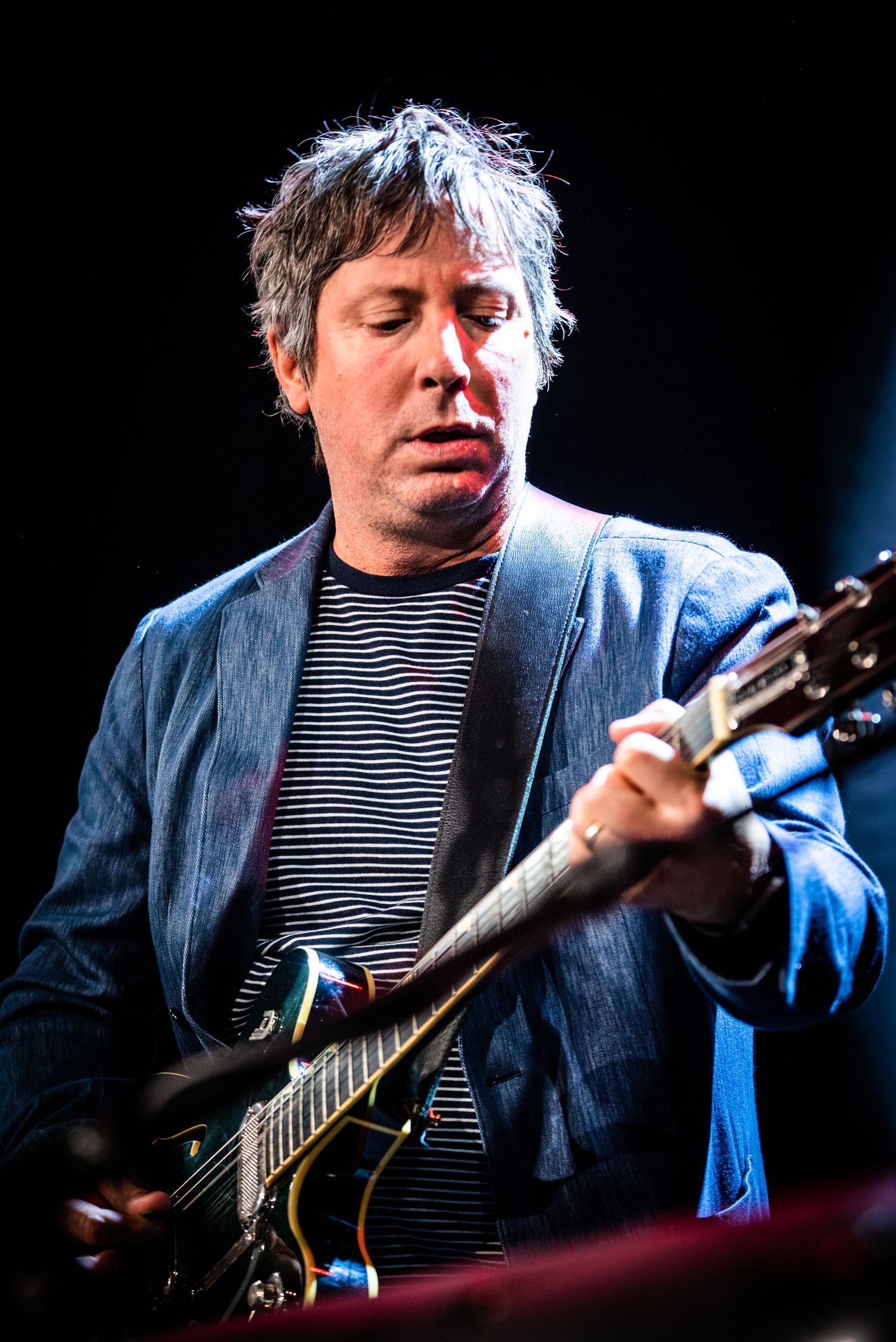 Musician in striped shirt and blazer plays guitar during intimate concert performance.