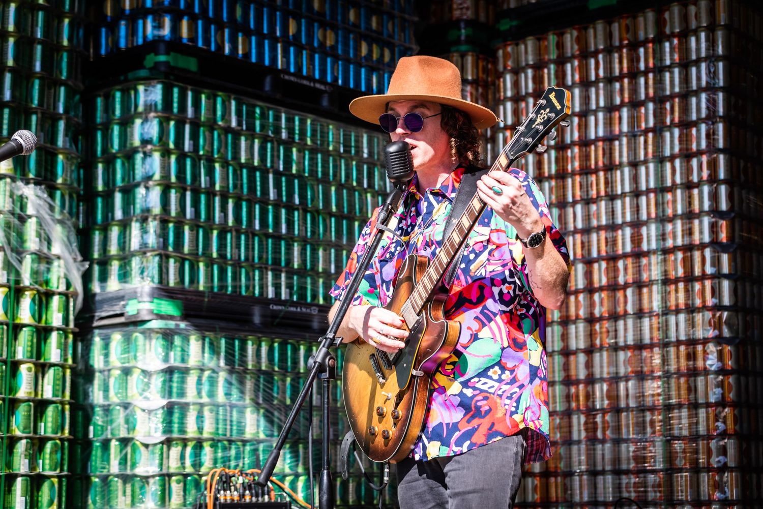 Musician performing with a guitar while wearing a colorful Hawaiian shirt and brown hat.