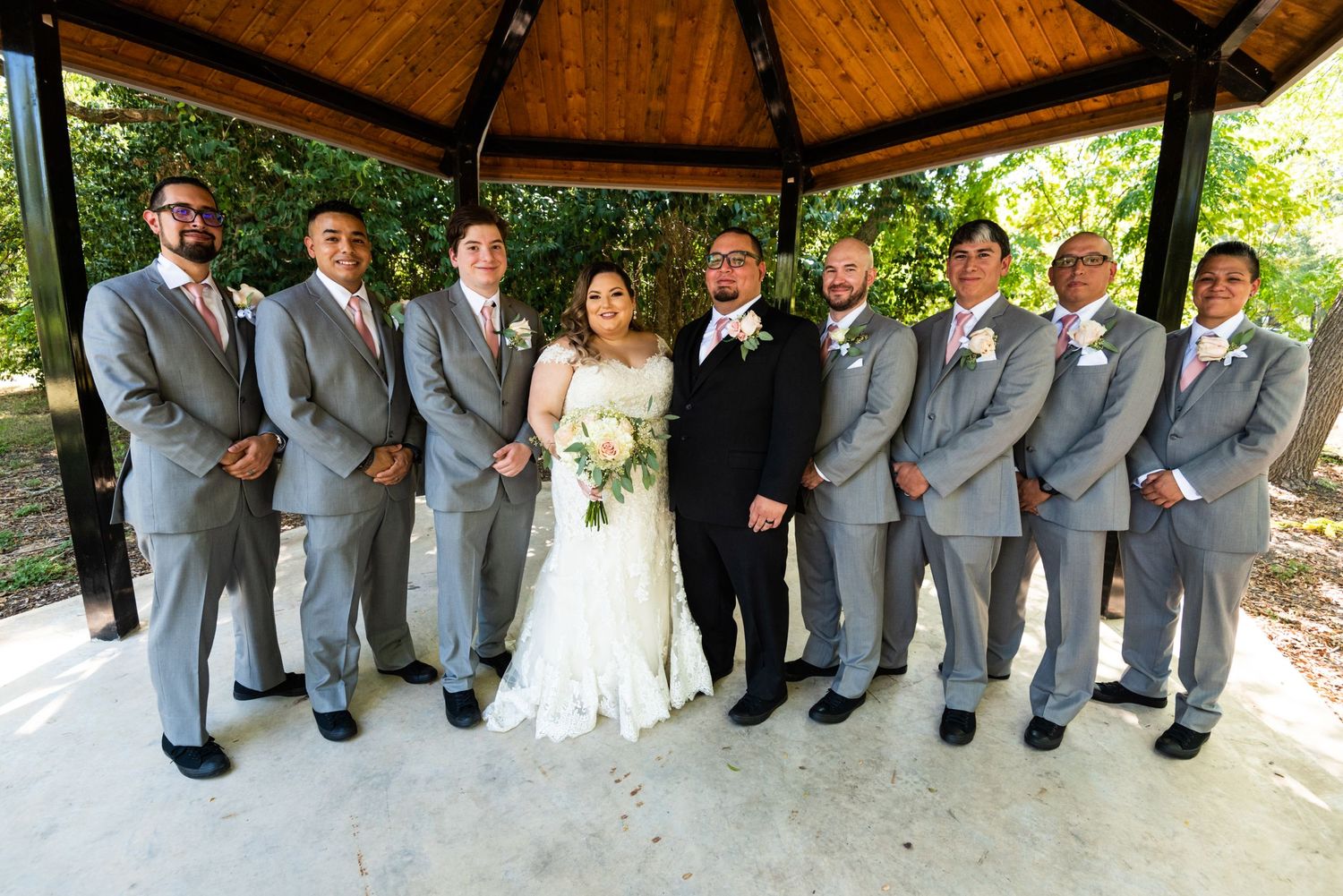 Wedding party in grey suits poses with couple under wooden gazebo outdoors.