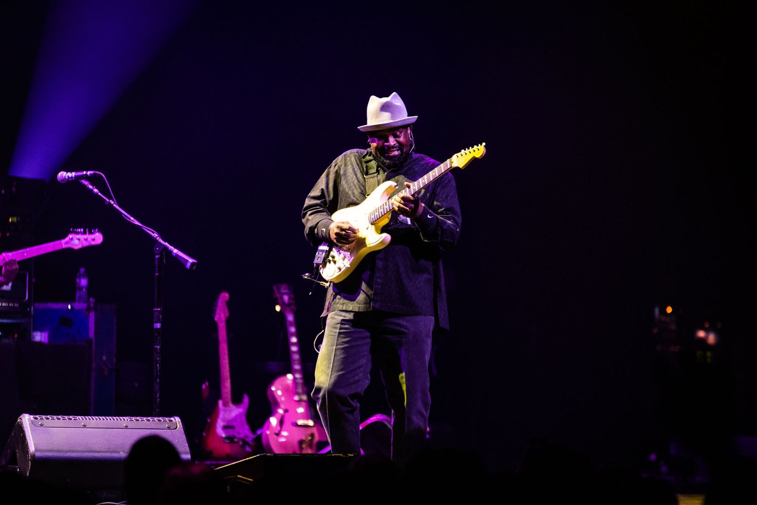 A guitarist performs on stage in a dark concert setting with purple lighting and musical equipment visible.