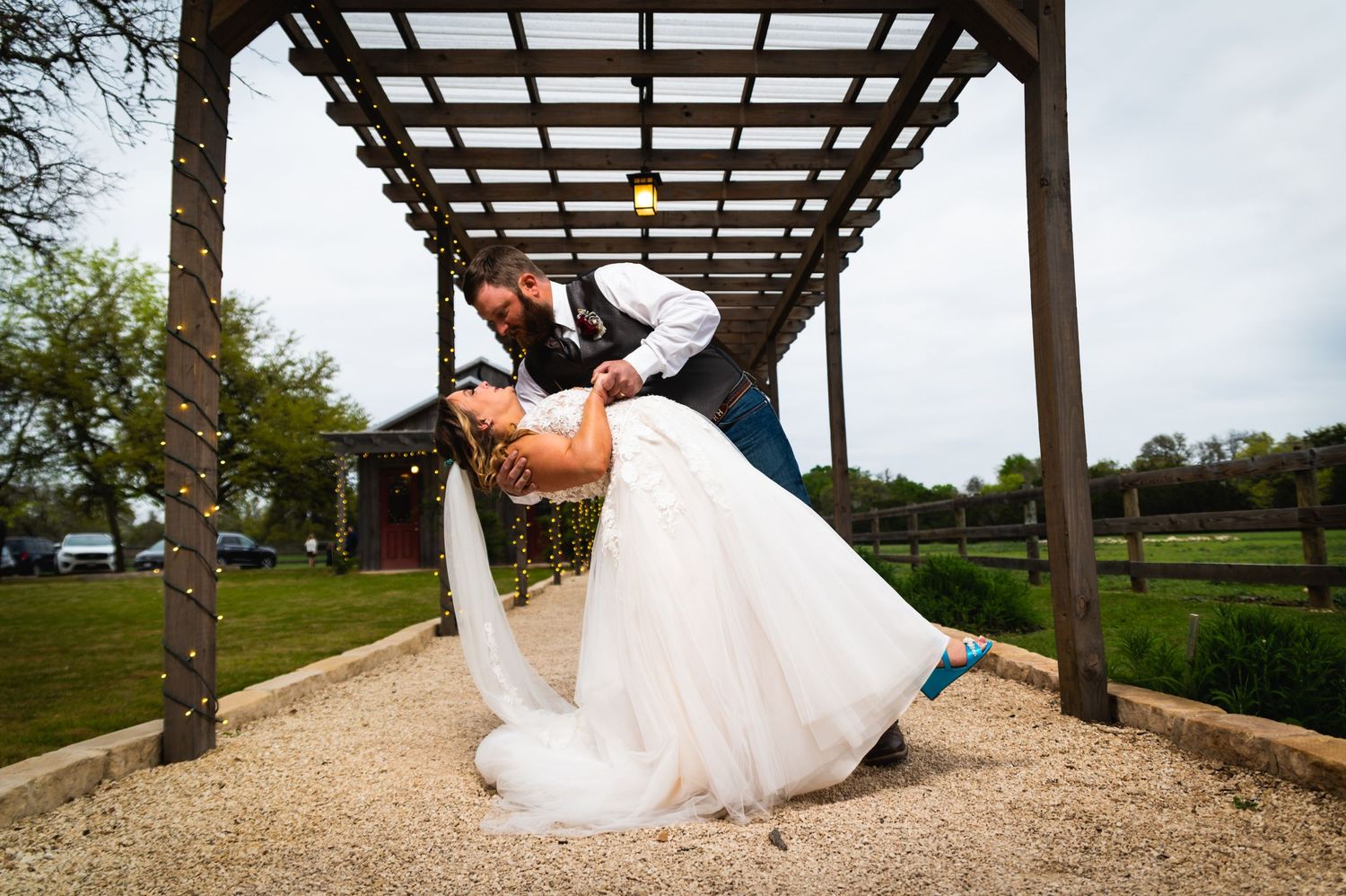 Romantic dip kiss between couple under wooden pergola at rustic outdoor venue.