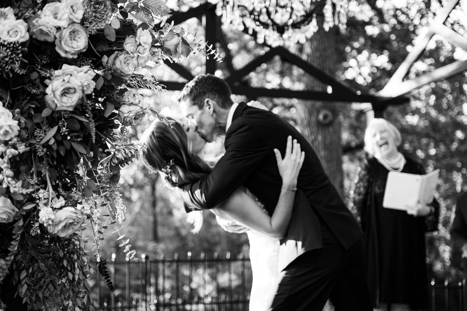 A black and white romantic dip kiss between newlyweds under a flower-adorned arch.