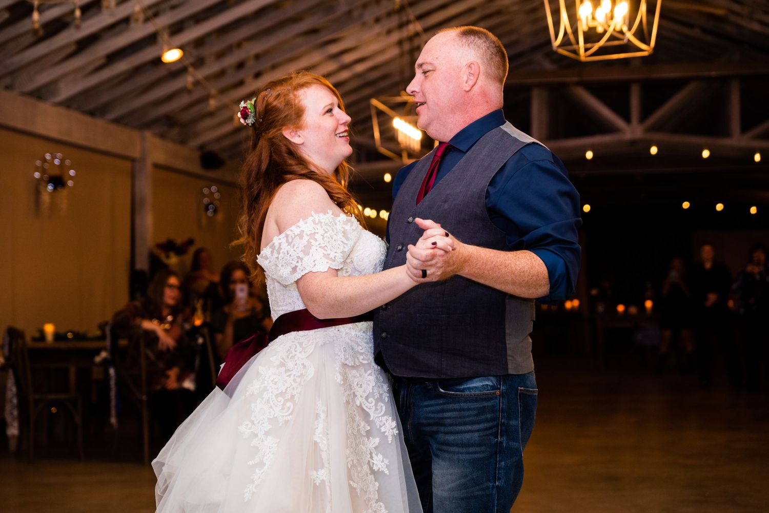 A wedding couple dances together in a rustic venue with string lights overhead.