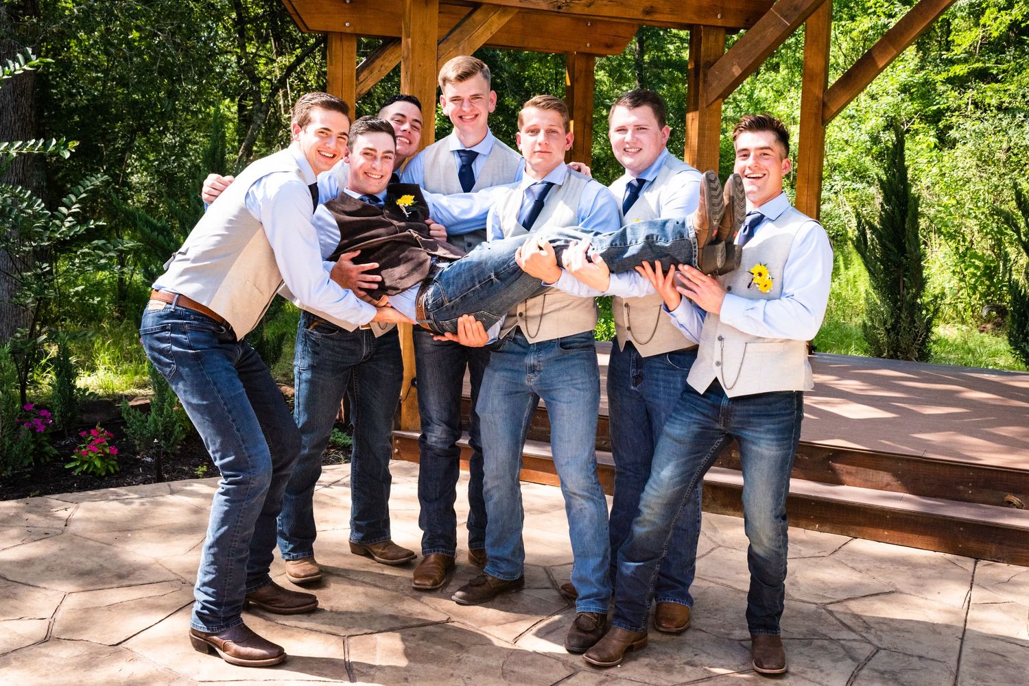 Groomsmen in blue shirts and vests playfully pose together under outdoor pavilion.