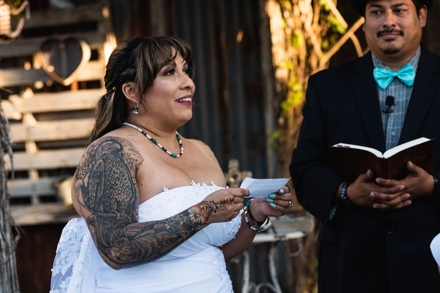 A tattooed bride in a white dress reads her vows during an outdoor ceremony.