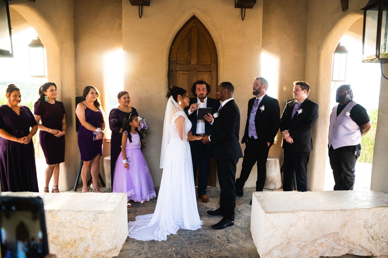 Wedding ceremony under arched colonnade with bridal party in purple dresses watching couple exchange vows.