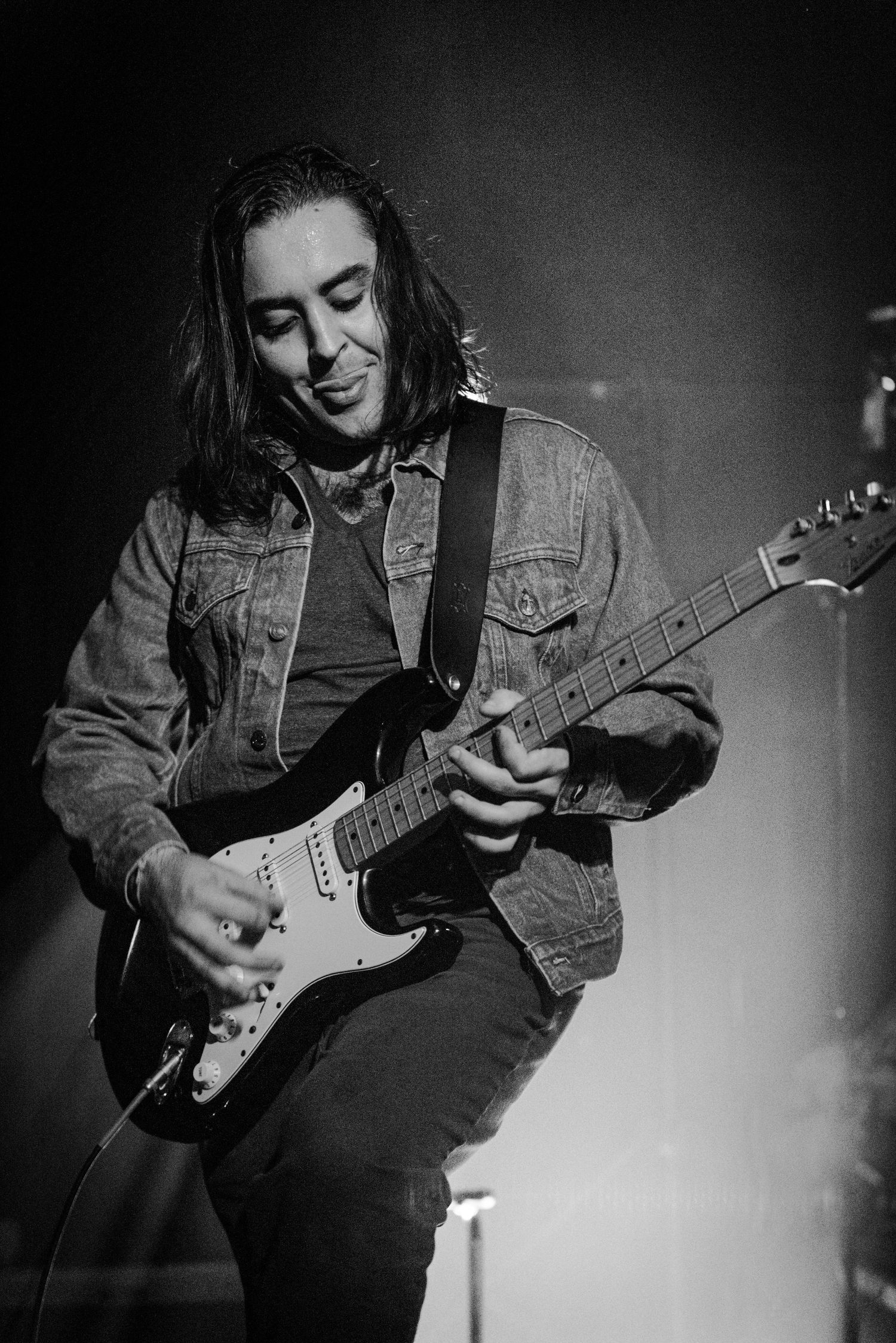 A guitarist in denim jacket plays electric guitar on a dimly lit stage.