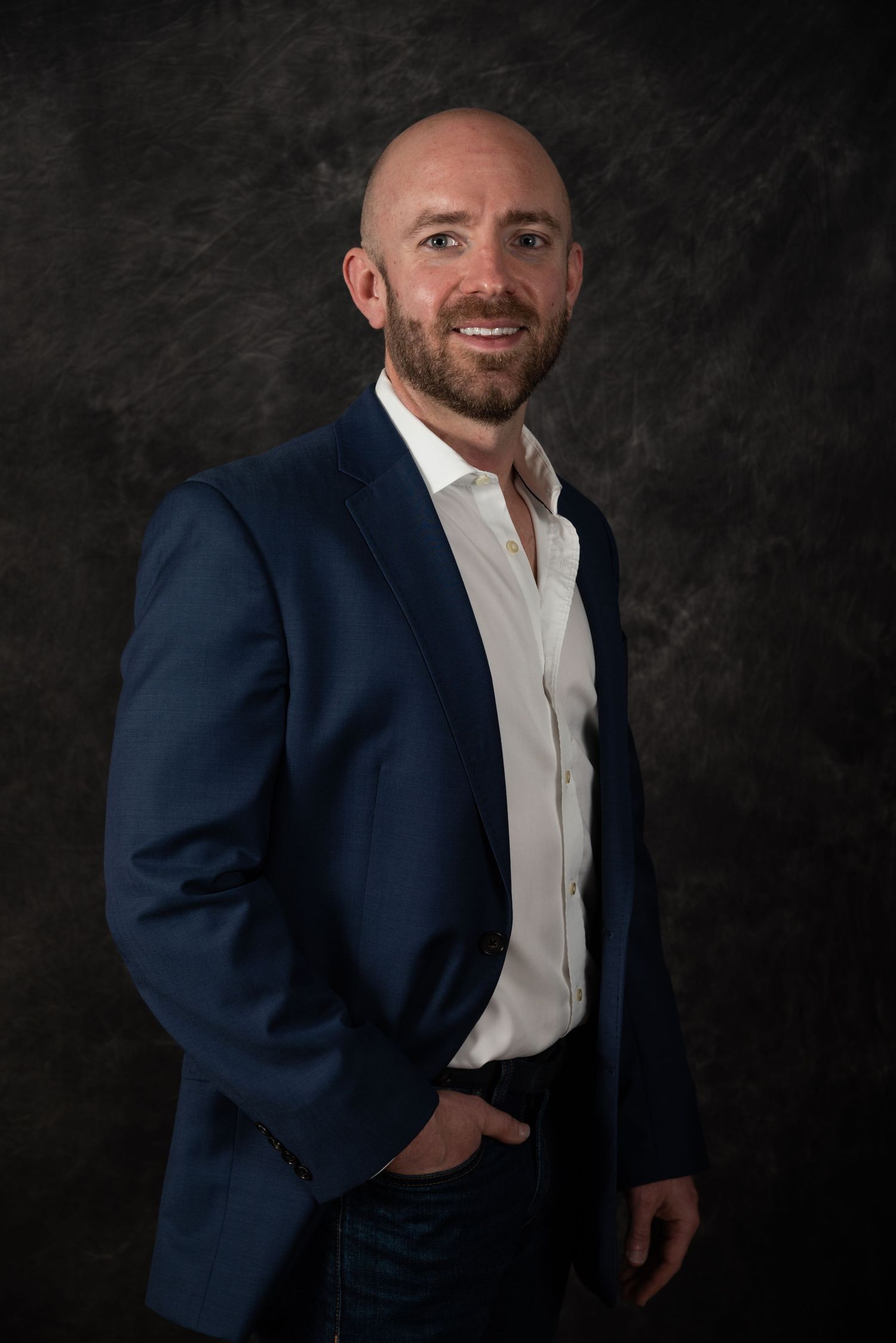 Professional headshot of person in navy blazer and white shirt against dark background.