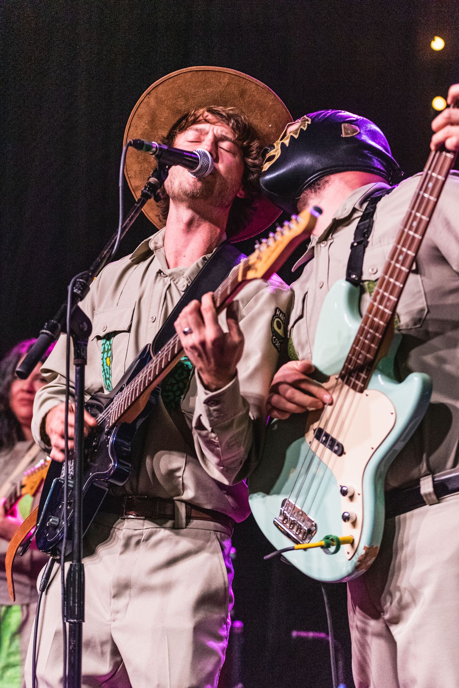 Musicians in matching outfits perform together on stage with vibrant purple lighting.