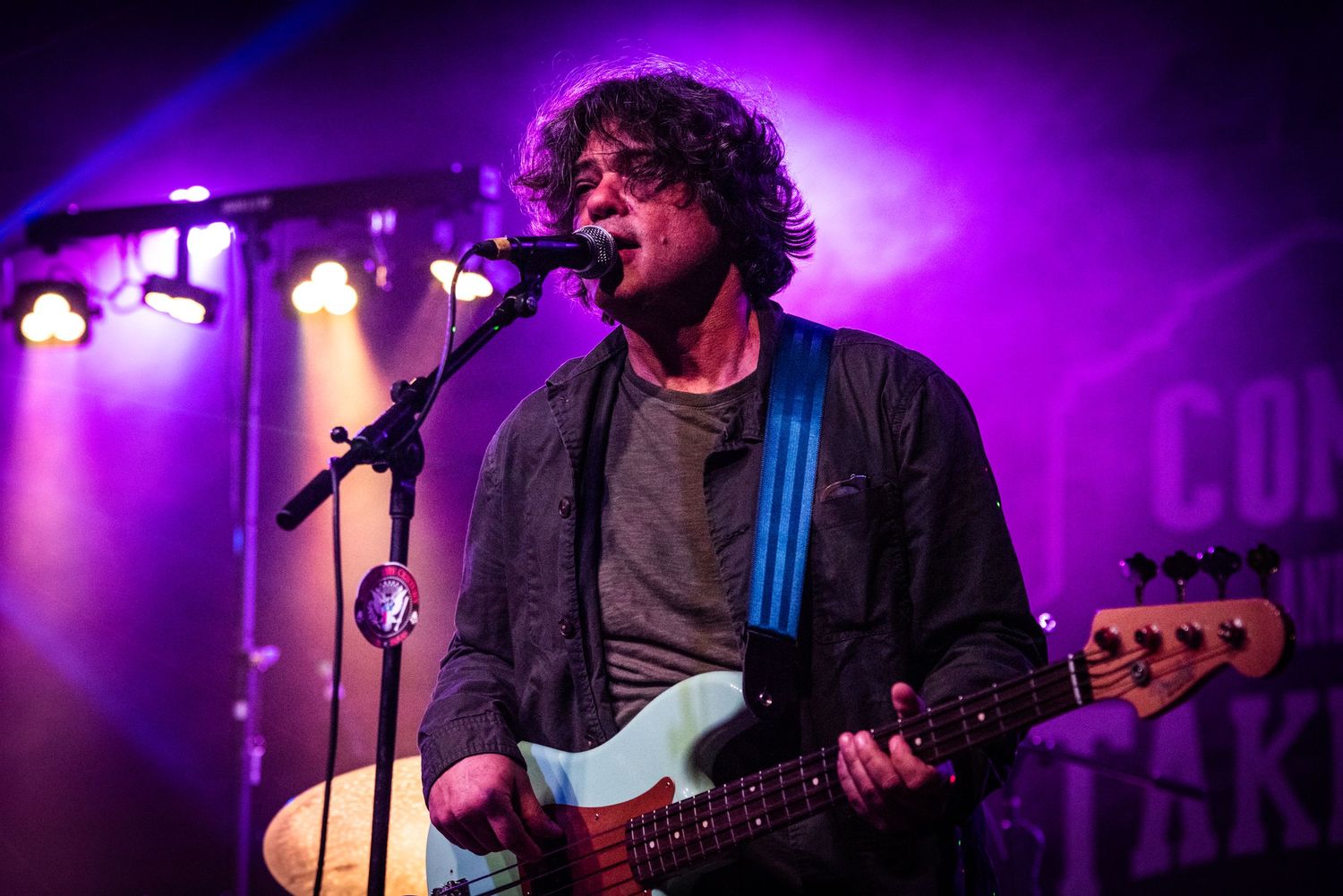 A performer plays bass guitar and sings under purple stage lighting during a concert.